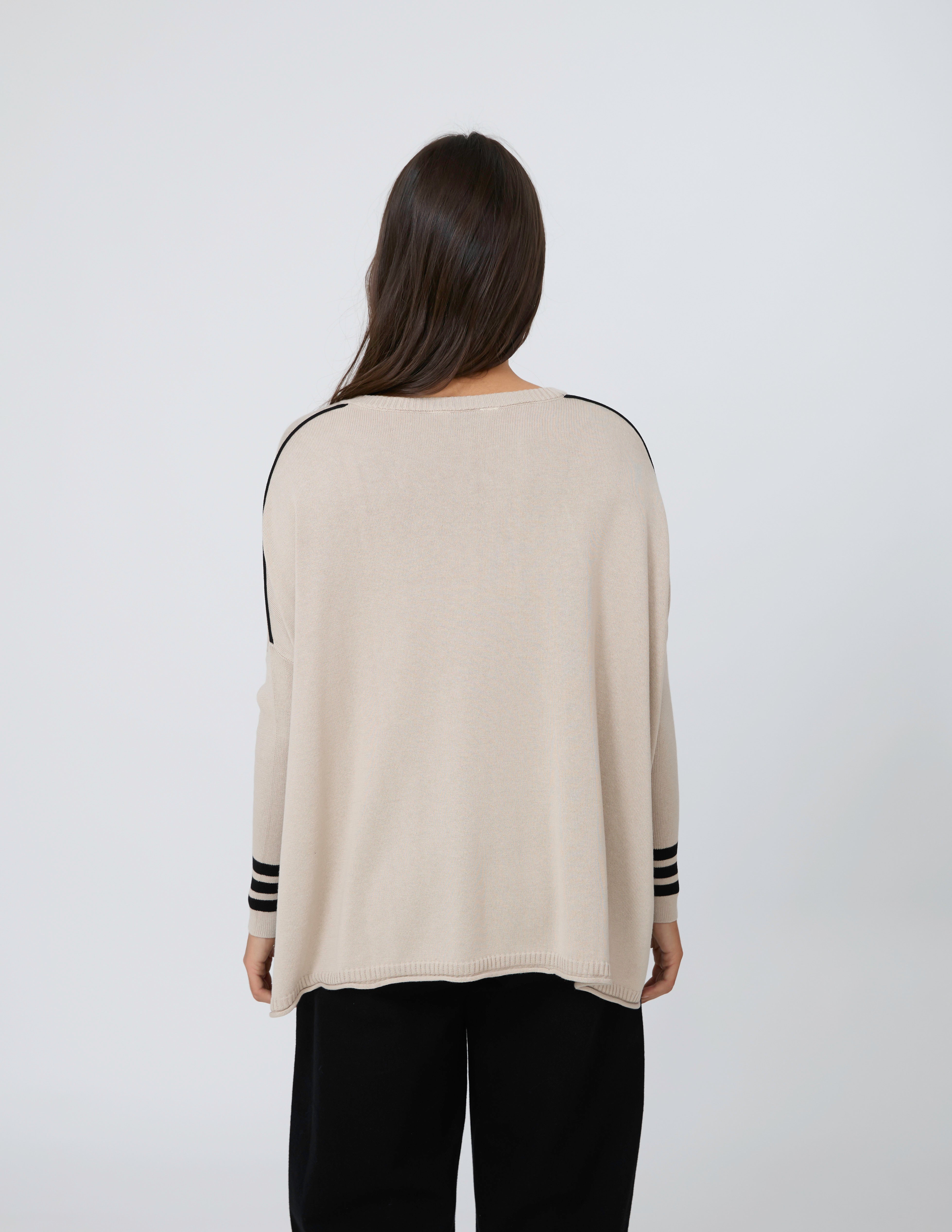 A woman viewed from behind wearing a light beige knit sweater with black stripe details on the shoulders and cuffs, paired with black pants, against a plain light gray background.