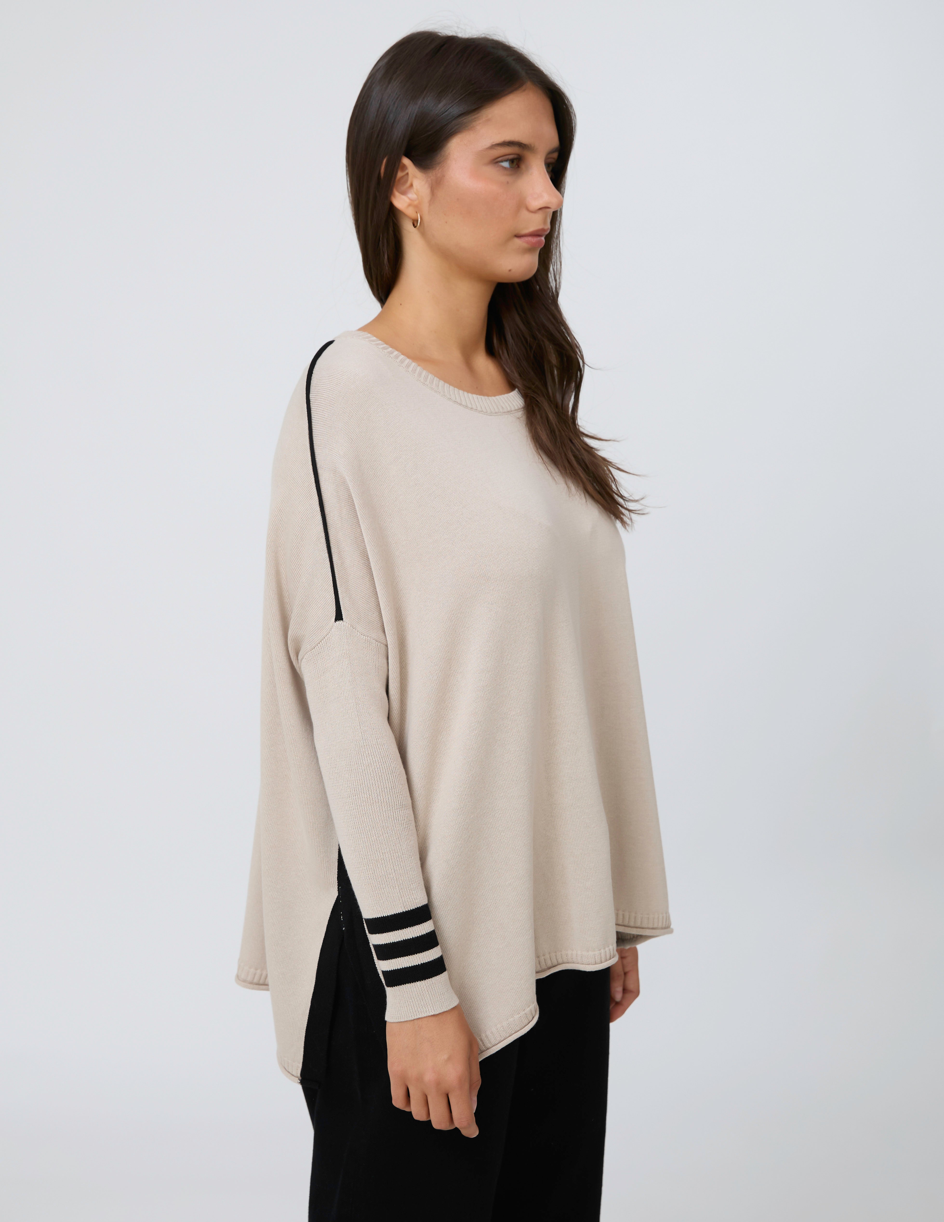 A woman modeling a light beige knit sweater with black stripe details on the sleeve cuffs and a black accent line on the shoulder, paired with black pants, against a plain light gray background.