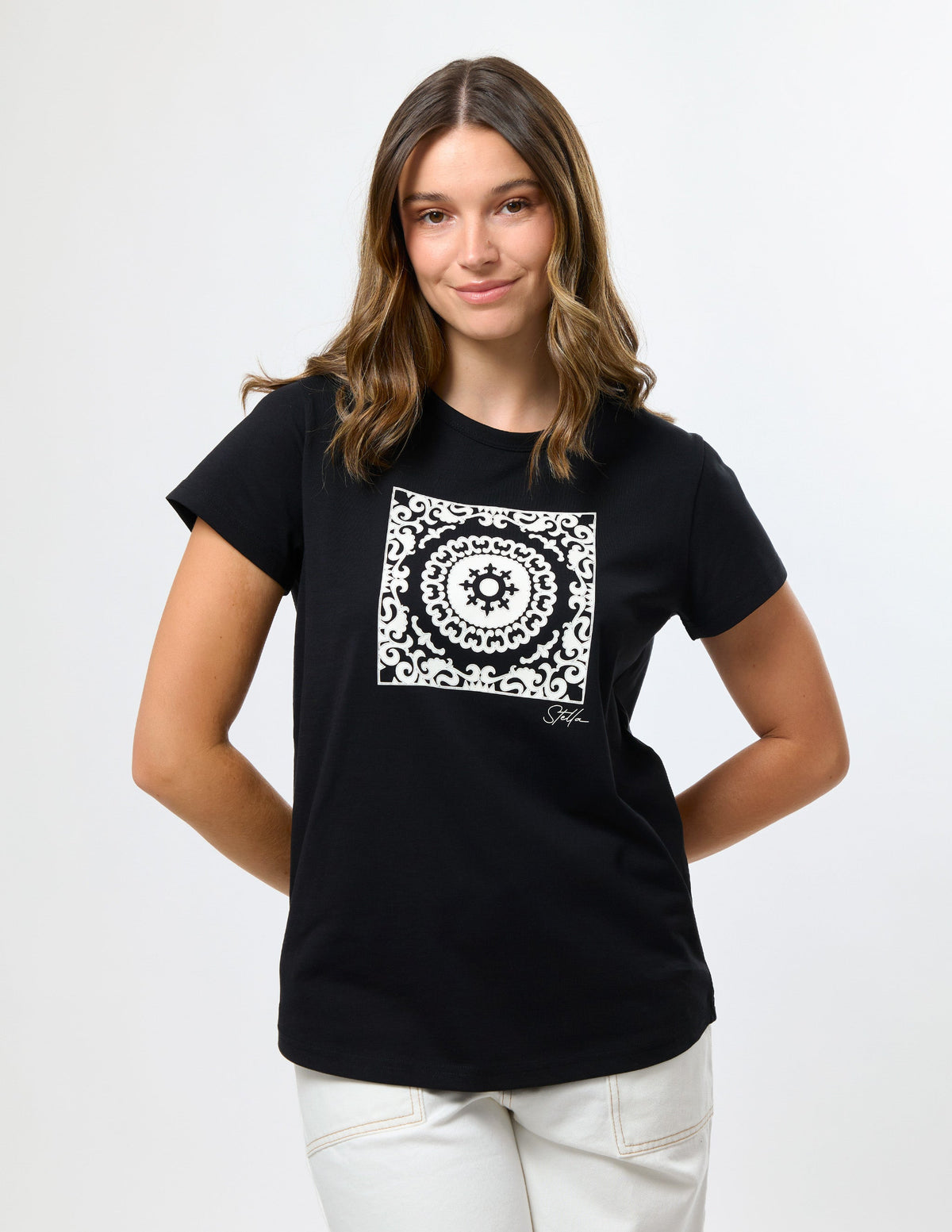 A woman modeling a black short-sleeve t-shirt with a white square mandala graphic on the chest, paired with white pants, against a plain light gray background.