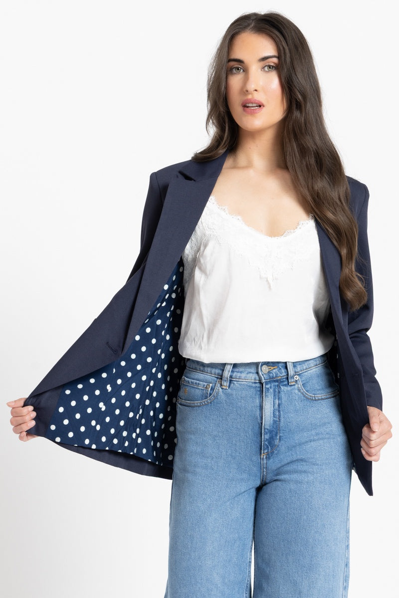 Woman wearing a navy blazer with a polka dot lining, white top, and blue jeans on a white background