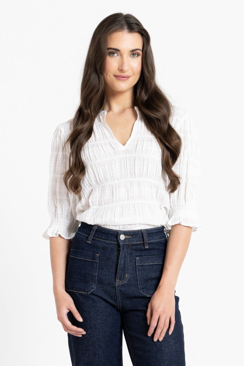 A woman with long wavy brown hair wearing a white textured blouse with ruffled sleeves and dark blue jeans, standing against a plain white studio background.