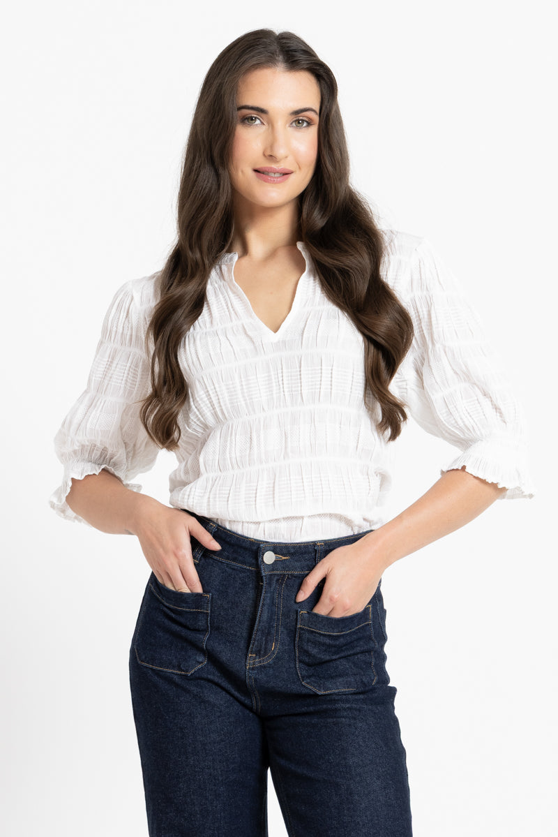 A woman modeling a white textured blouse with ruffled sleeves and dark blue jeans against a plain white studio background.