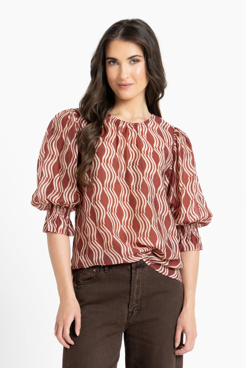 A woman modeling a rust-red and cream wavy patterned blouse with puff sleeves and a gathered waist, paired with dark brown pants, against a plain white background.