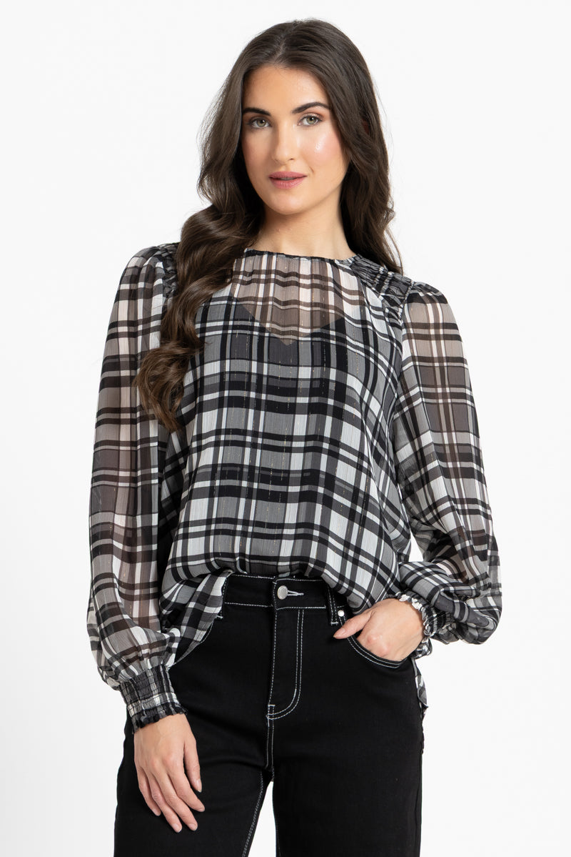 A woman wearing a black, white, and gray plaid blouse with sheer long sleeves and puff sleeves, paired with black pants, against a plain white studio background.