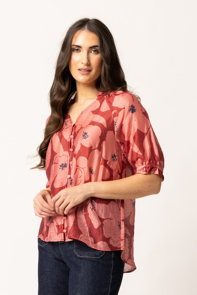 A woman modeling a short-sleeve button-up blouse with a pink and red floral pattern, paired with dark blue jeans, against a plain white studio background.