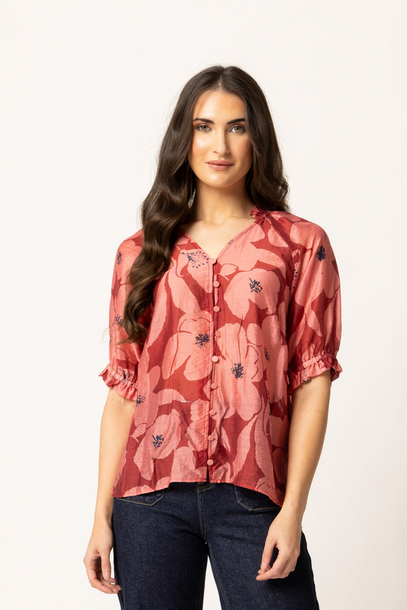 A woman modeling a short-sleeve button-up blouse with a dusty rose and coral floral print, paired with dark blue jeans, against a plain white studio background.