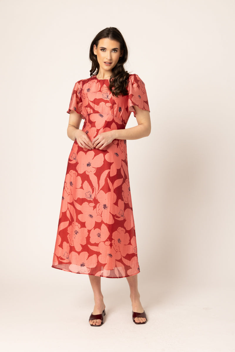A woman modeling a midi-length red dress with a large pink floral pattern, short puff sleeves, and a round neckline, set against a plain light beige studio background.