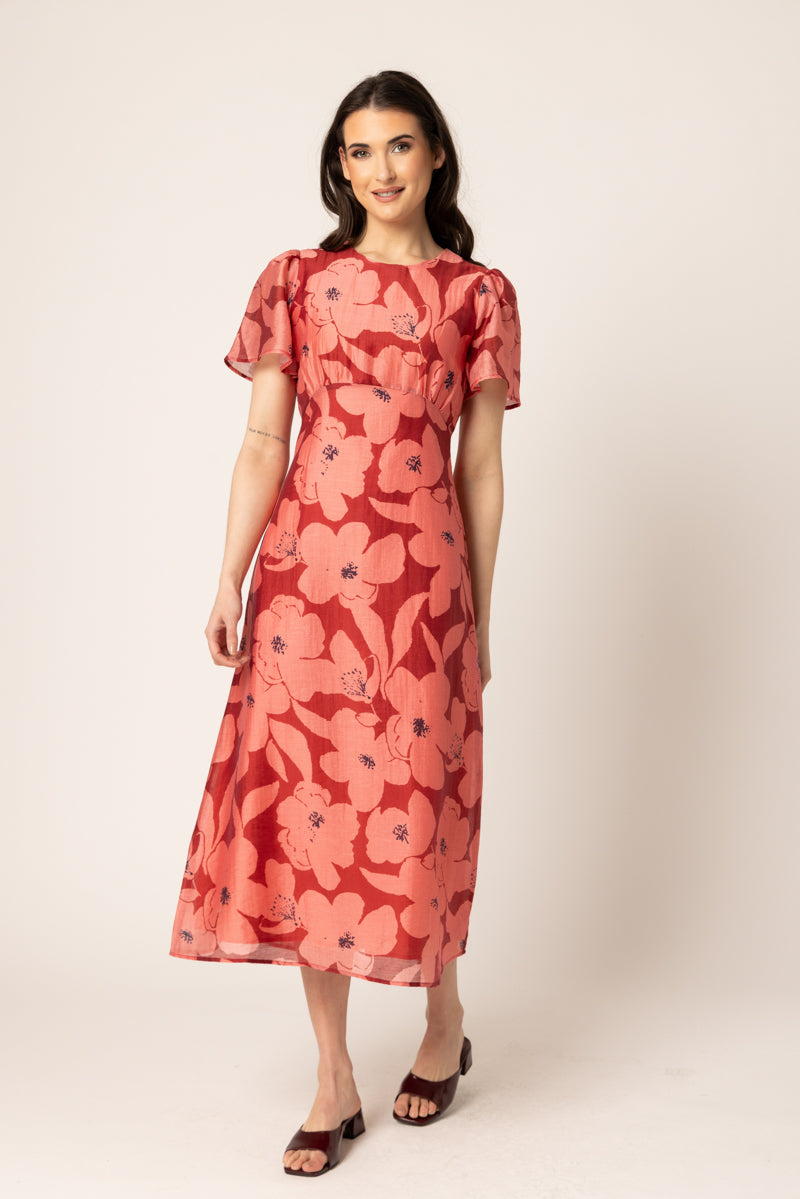 A woman modeling a midi-length coral pink dress with a large floral print, short puff sleeves, and a round neckline, paired with dark brown heeled sandals against a plain beige studio background.