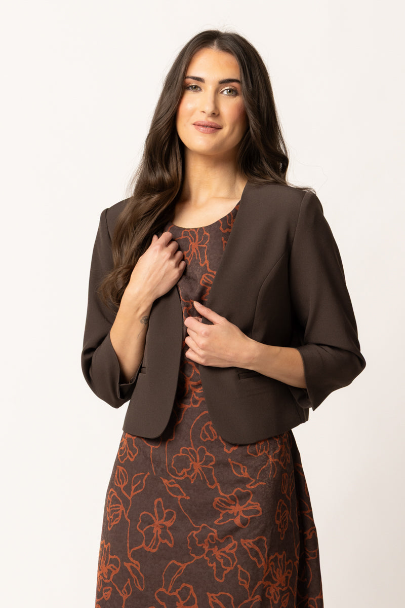 A woman modeling a dark brown cropped jacket over a brown and rust floral dress, photographed against a clean off-white studio background.