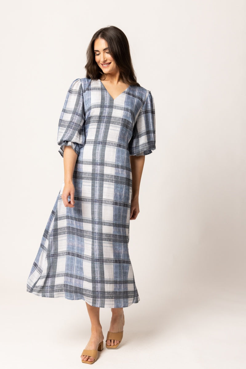A woman modeling a blue and white plaid midi dress with puff sleeves and a V-neckline, standing against a plain light beige studio background.