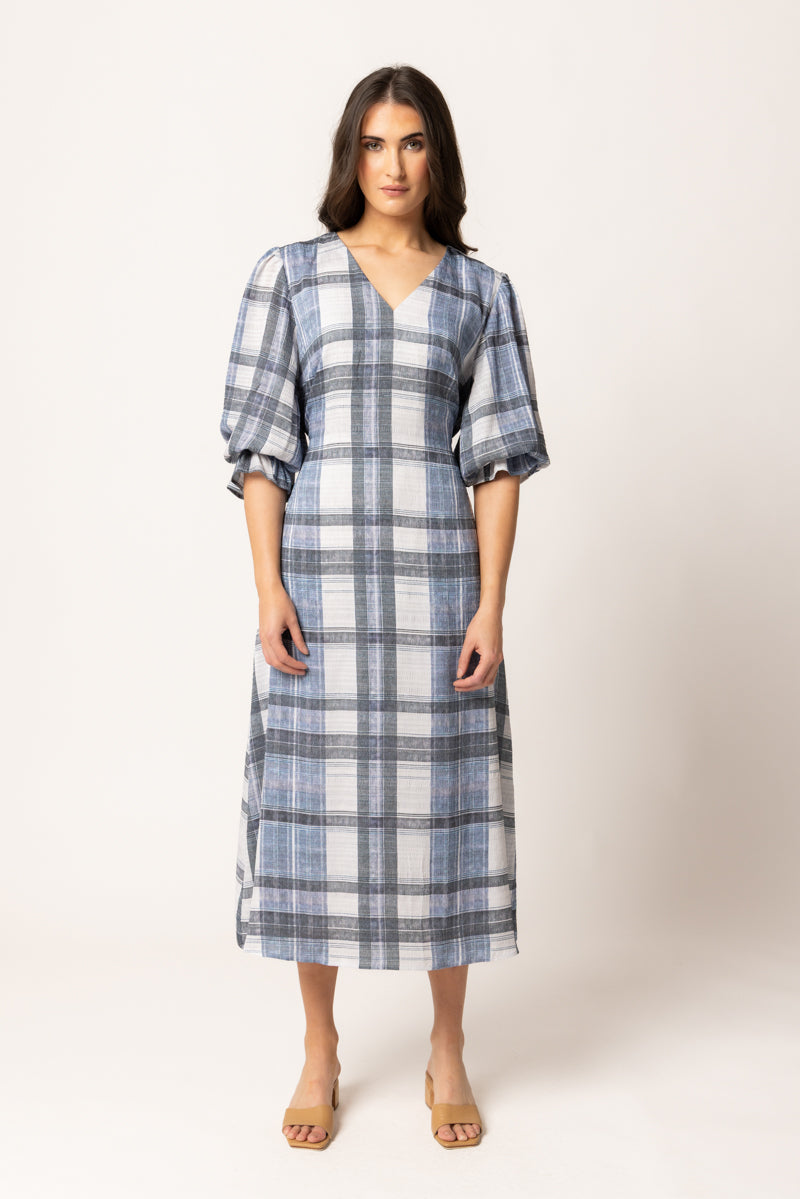 A woman modeling a blue and white plaid midi dress with puff sleeves and a V-neckline, standing against a plain light beige studio background.