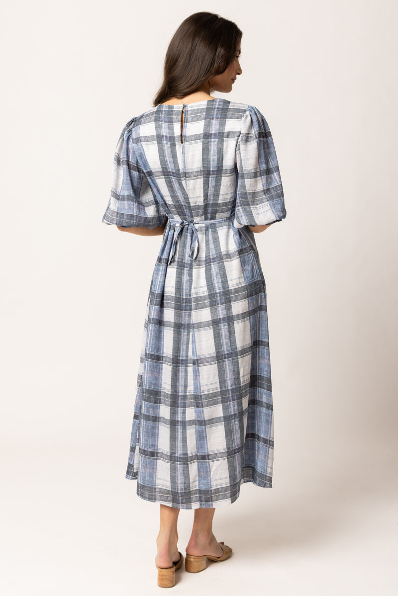 A woman viewed from the back wearing a midi dress with a blue, gray, and white plaid pattern, puff sleeves, and a tie waist, paired with tan flat sandals against a light beige studio background.