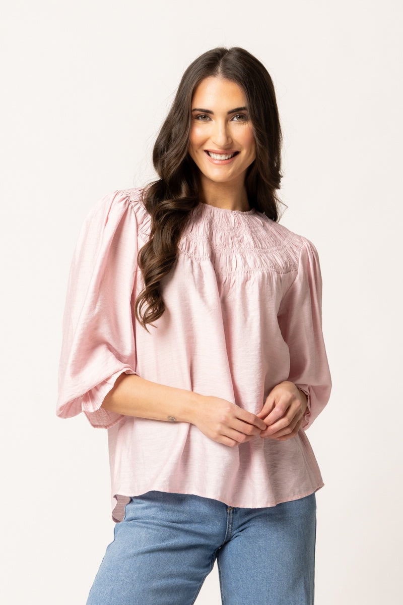A woman with long dark hair wearing a light pink smocked blouse with puff sleeves and blue jeans, standing against a plain white studio background.