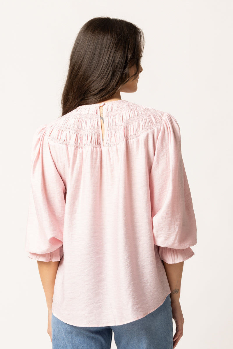 A woman viewed from the back wearing a light pink blouse with voluminous elbow-length puff sleeves, a smocked neckline, and a small gold button closure at the back center. She is also wearing blue jeans against a plain off-white studio background.