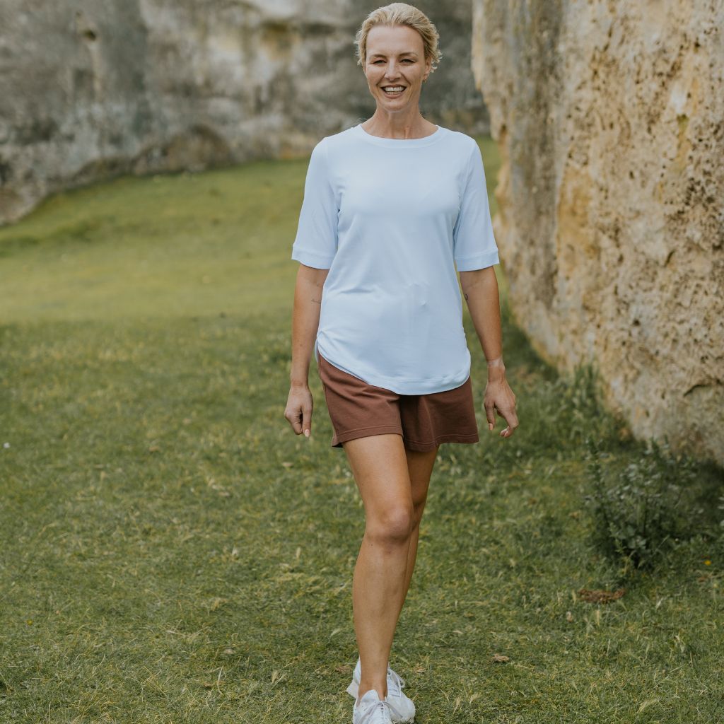 A smiling woman walking outdoors on a grassy area next to a large rock wall, wearing a light blue short-sleeve crewneck t-shirt, brown shorts, and white sneakers.