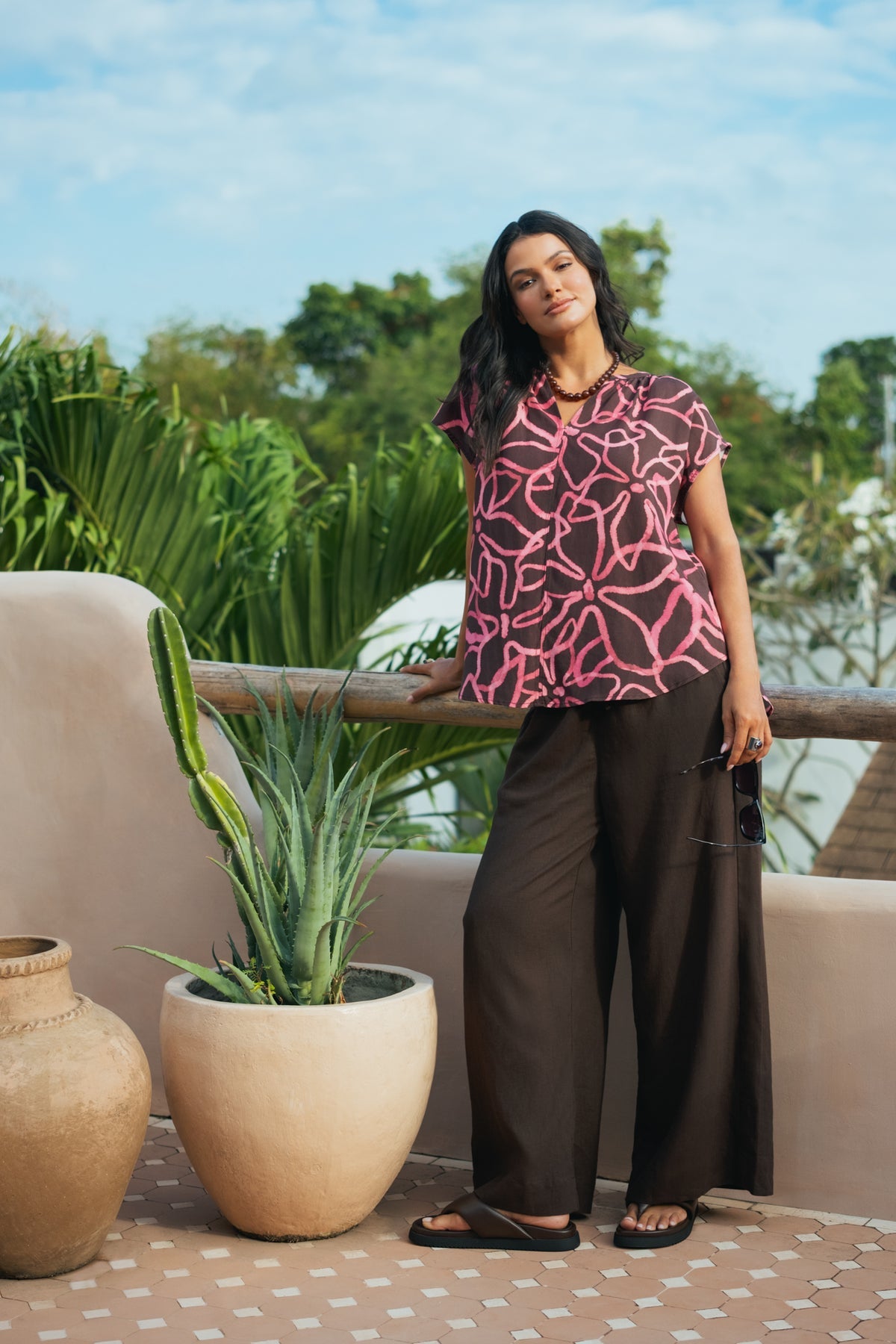 A woman stands on a balcony wearing a short-sleeve button-up blouse with a pink and black abstract pattern, paired with dark wide-leg pants and black sandals. The background features a beige wall, a wooden railing, potted plants, and a blue sky.