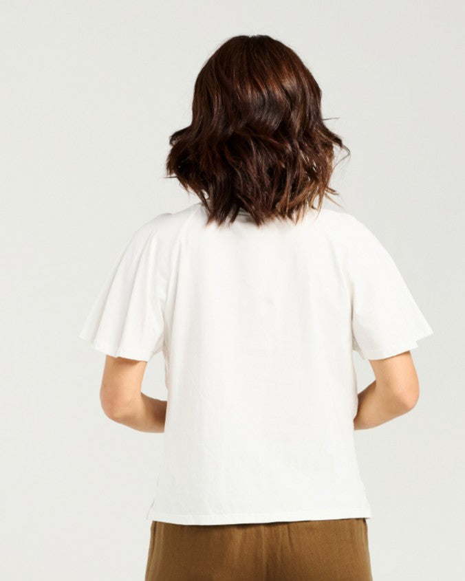 A woman viewed from behind wearing a white short-sleeve t-shirt and brown pants, set against a plain light gray studio background.