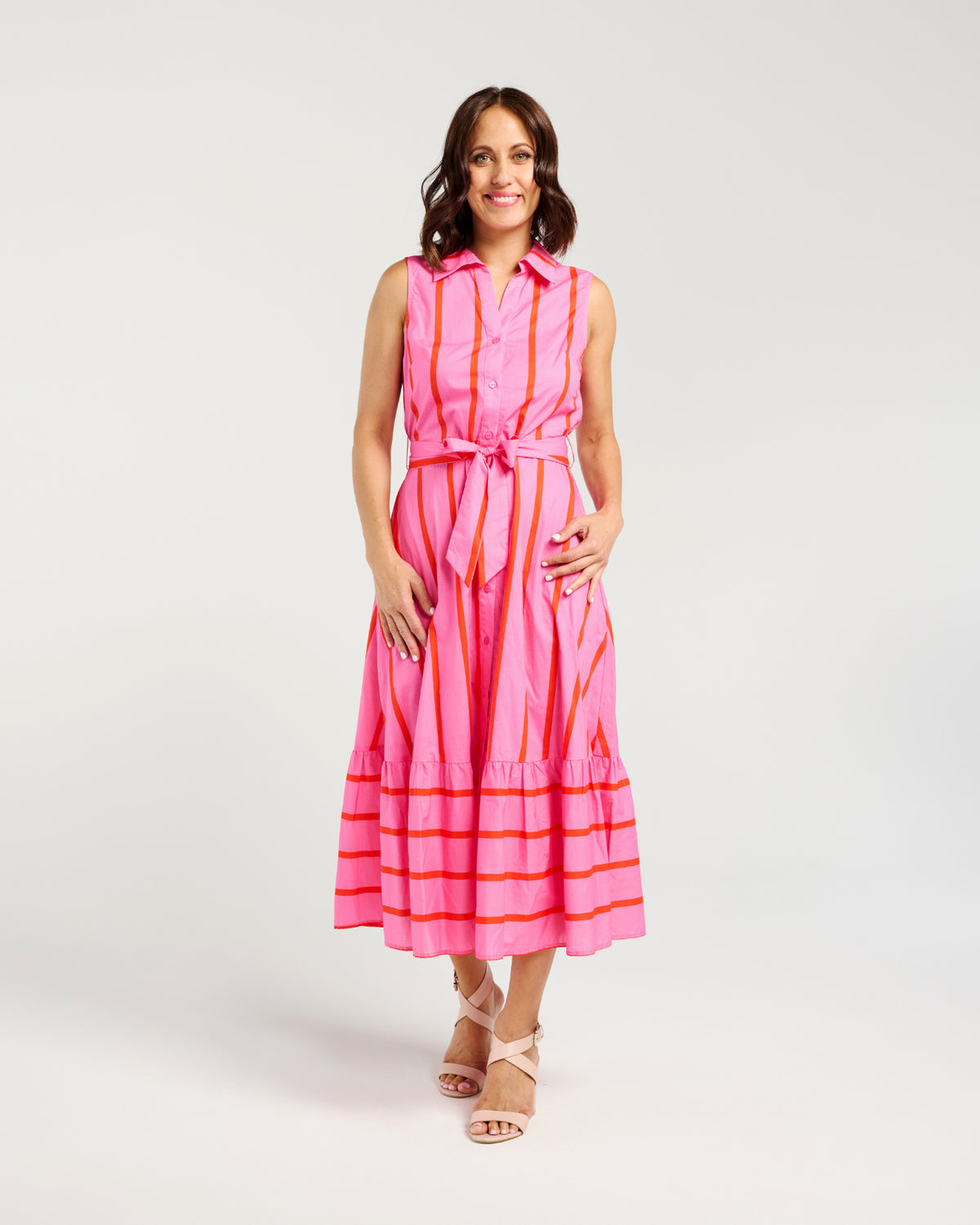 A woman modeling a sleeveless pink midi dress with vertical orange stripes, a matching tie belt at the waist, and horizontal orange stripes along the tiered hem, photographed against a light gray studio background.