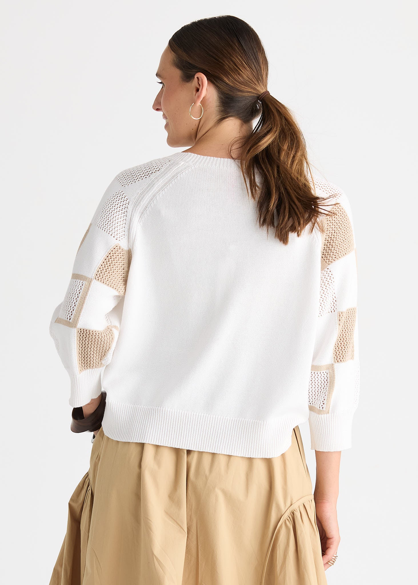 A woman shown from the back wearing a white knit sweater with beige open-knit panel details on the sleeves, paired with a beige skirt, against a light gray studio background.