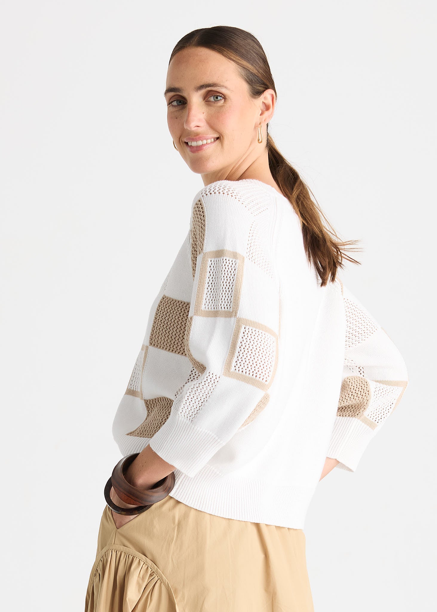 A woman wearing a white knit sweater with beige patchwork panels and sheer mesh details, paired with a beige skirt, photographed against a light gray studio background.