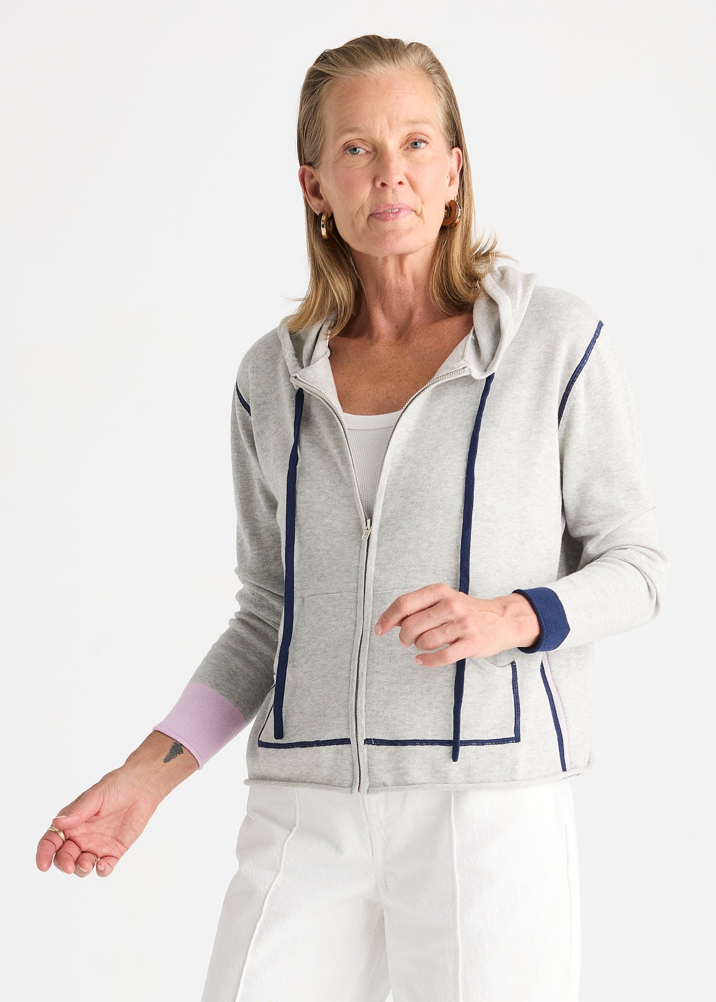 A woman wearing a light gray zip-up hoodie with navy blue contrast piping and cuffs, styled with white pants against a plain white studio background.