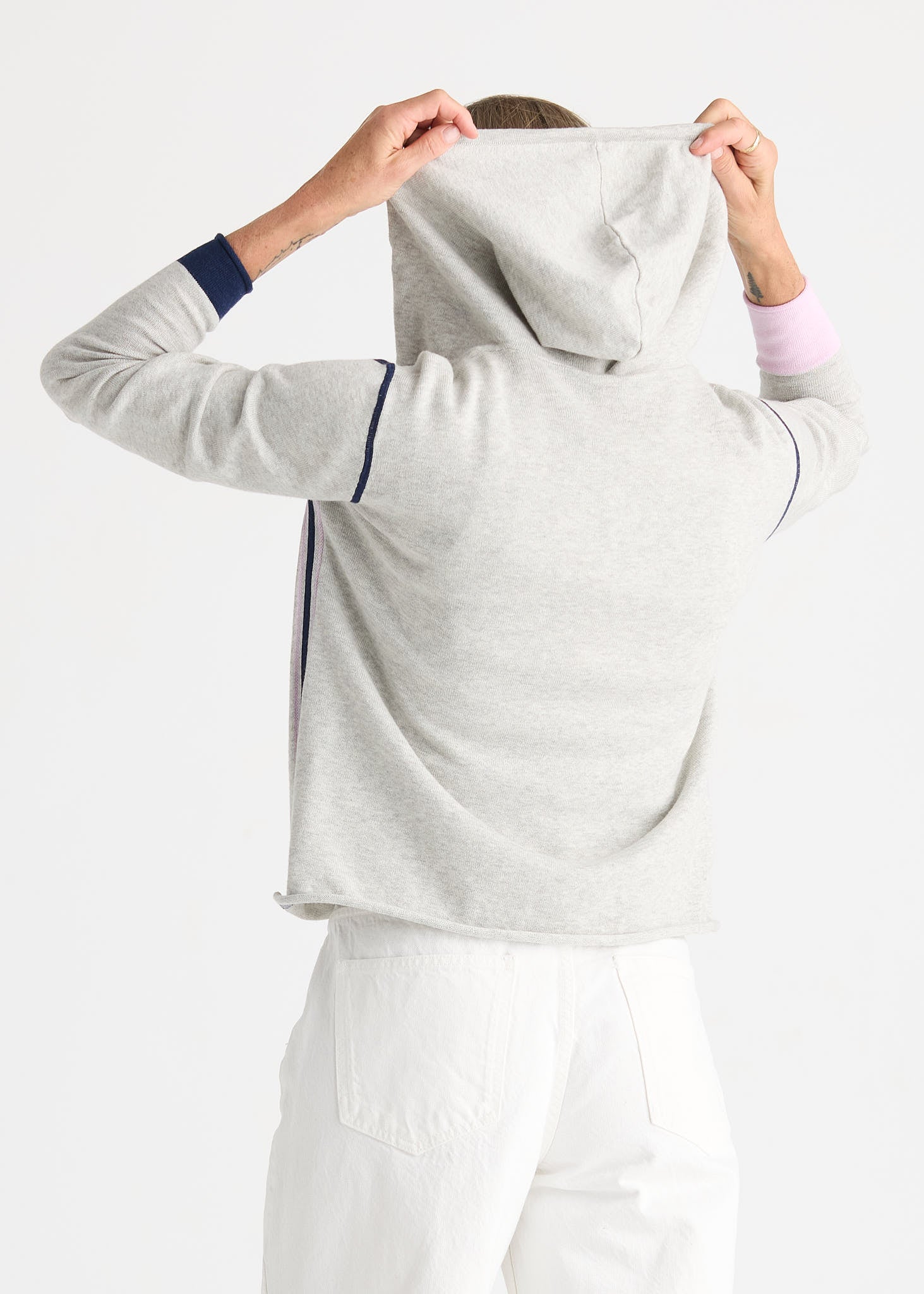 A person shown from the back wearing a light heather gray hooded sweatshirt with navy blue stripe accents on the sleeves and side, paired with white pants, against a plain white studio background.