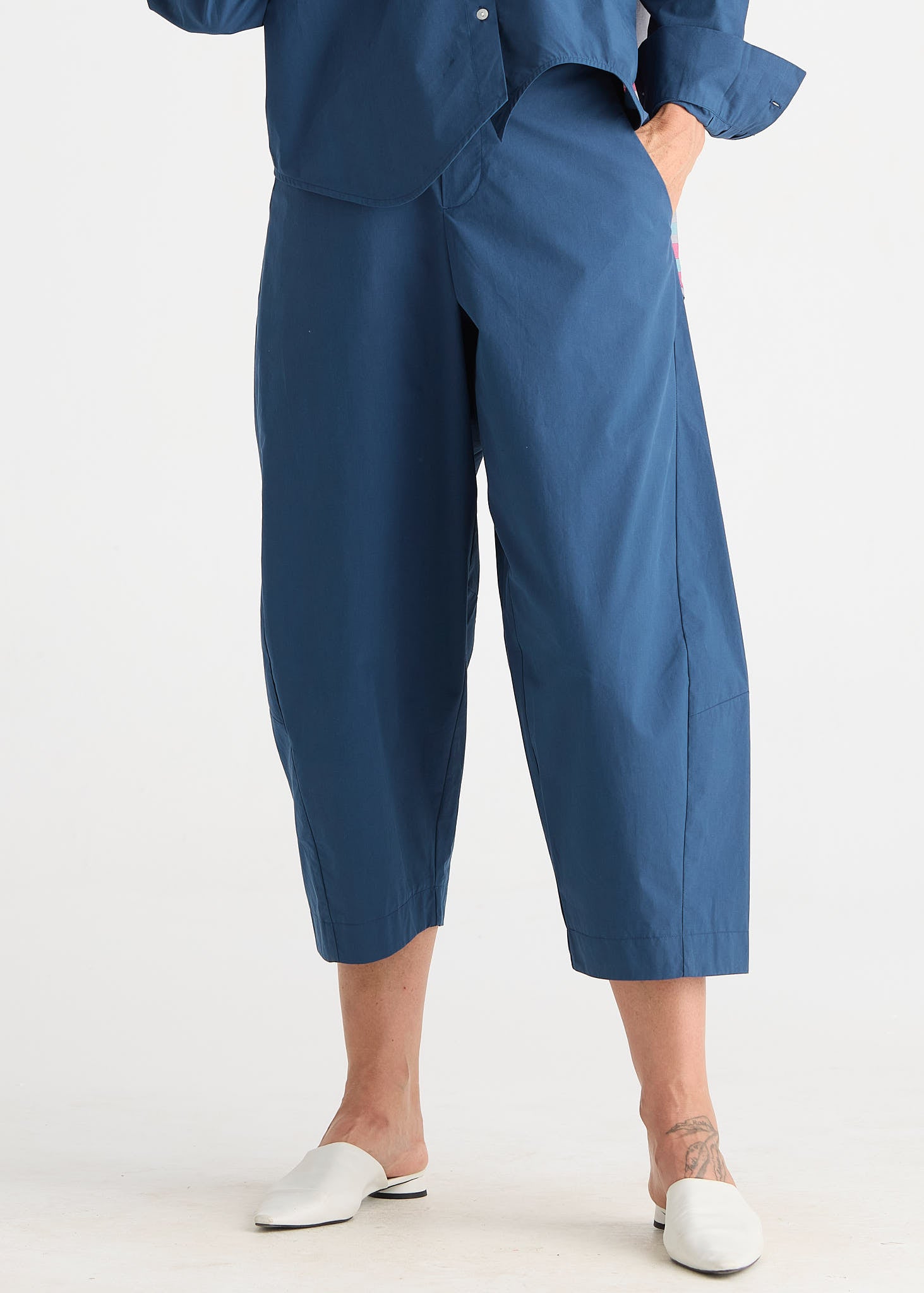 A person wearing loose, wide-leg cropped pants in a muted medium blue, paired with a matching blue top and white mule shoes, photographed against a plain light gray studio background.