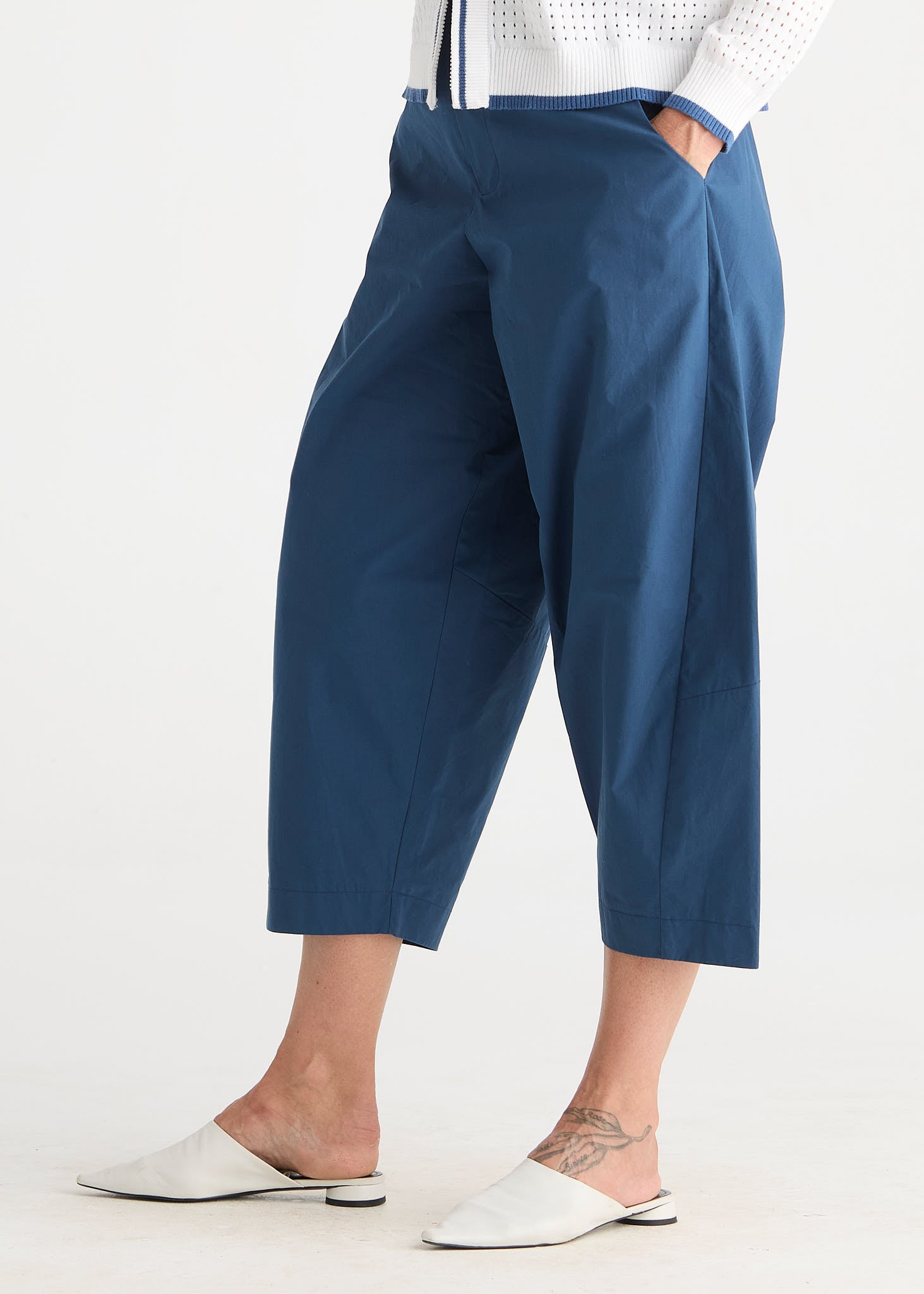 A person wearing loose, cropped blue pants with side pockets, paired with white mule flats and a white top, photographed against a plain light background.