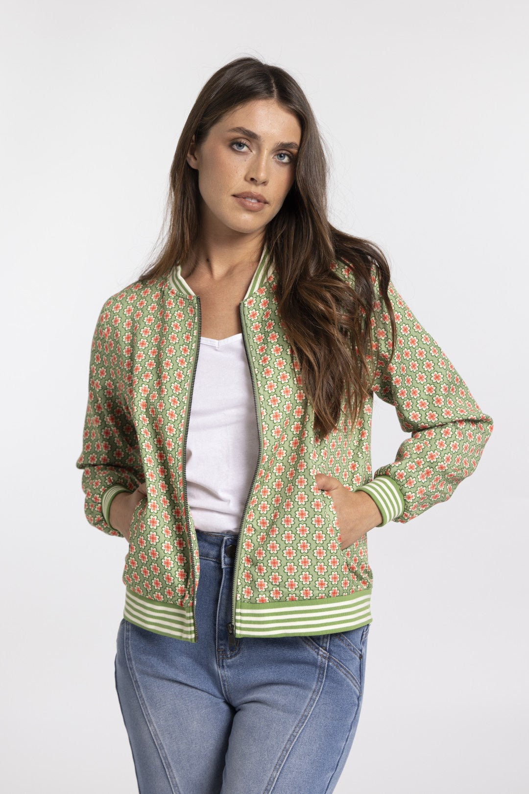 A woman modeling a green patterned bomber jacket with striped ribbed trim, worn over a white top and blue jeans, against a light gray studio background.