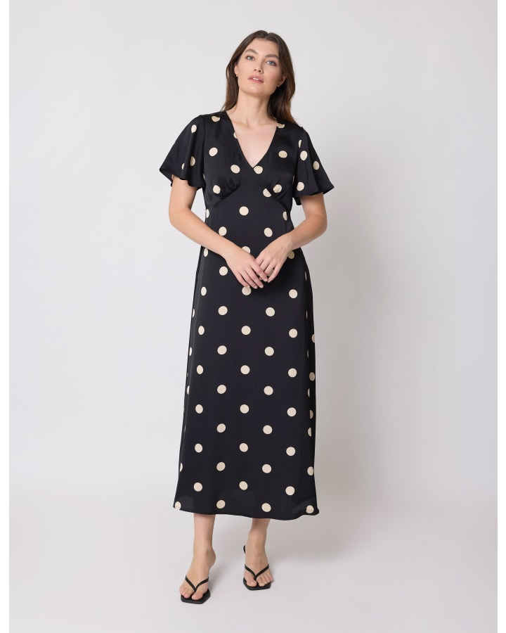 A woman modeling a black midi wrap dress with beige polka dots and flutter sleeves, standing against a light gray studio background.