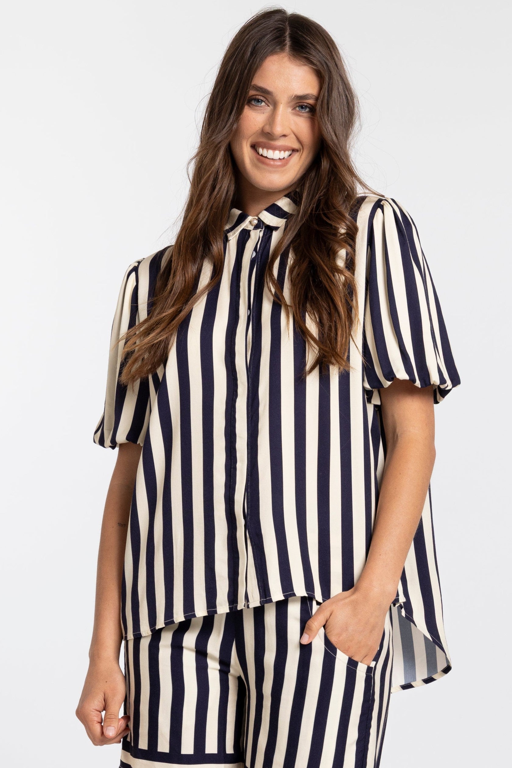 A woman modeling a matching two-piece set featuring a short-sleeve button-up blouse and shorts in a navy blue and cream vertical stripe pattern, set against a plain light gray background.
