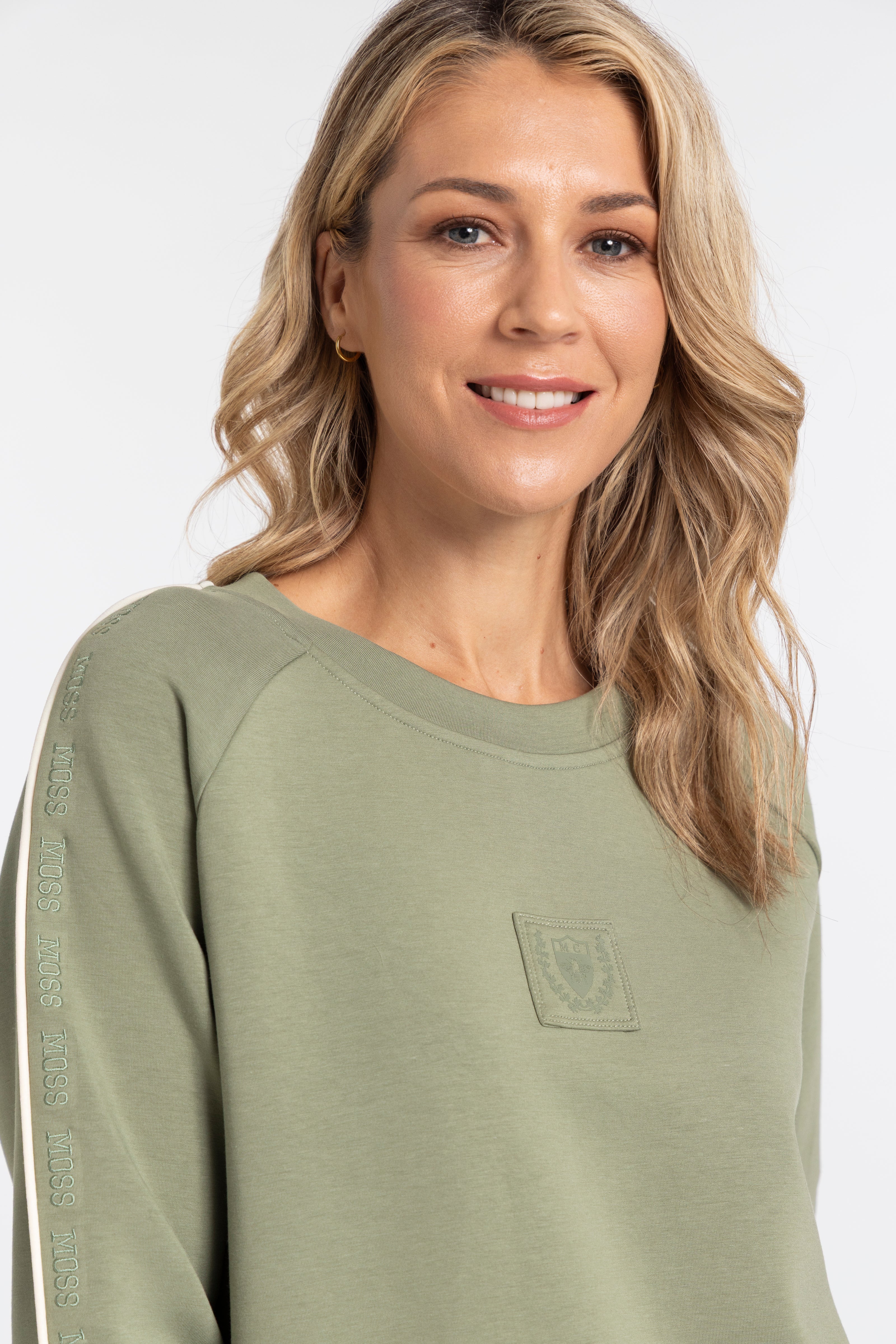 A woman with blonde hair wearing an olive green crewneck sweatshirt with a tonal chest patch and a subtle vertical logo print on the sleeve, set against a plain light gray background.
