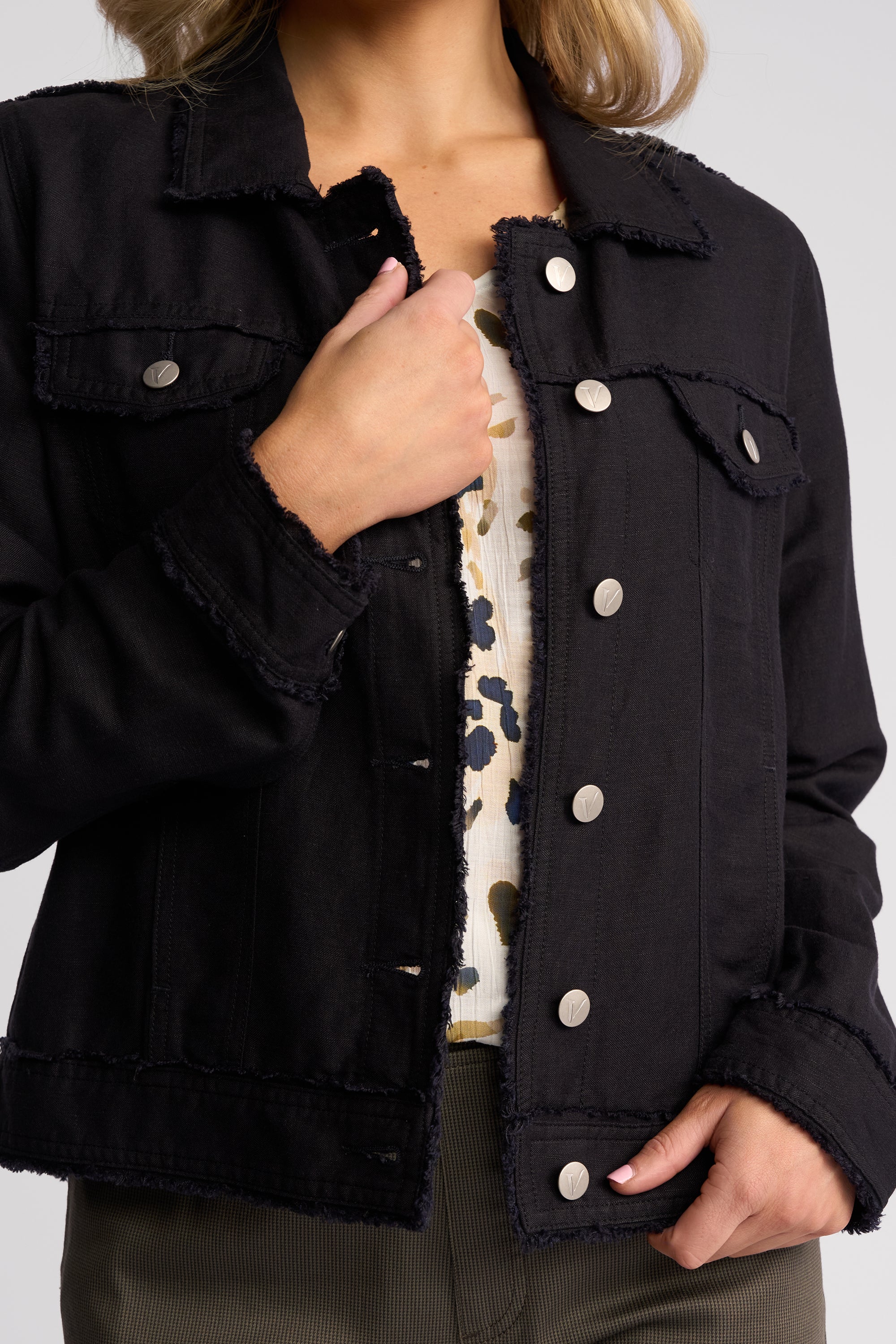 A close-up of a person wearing a black frayed denim jacket over a cream patterned top, with hands visible and a neutral studio background.