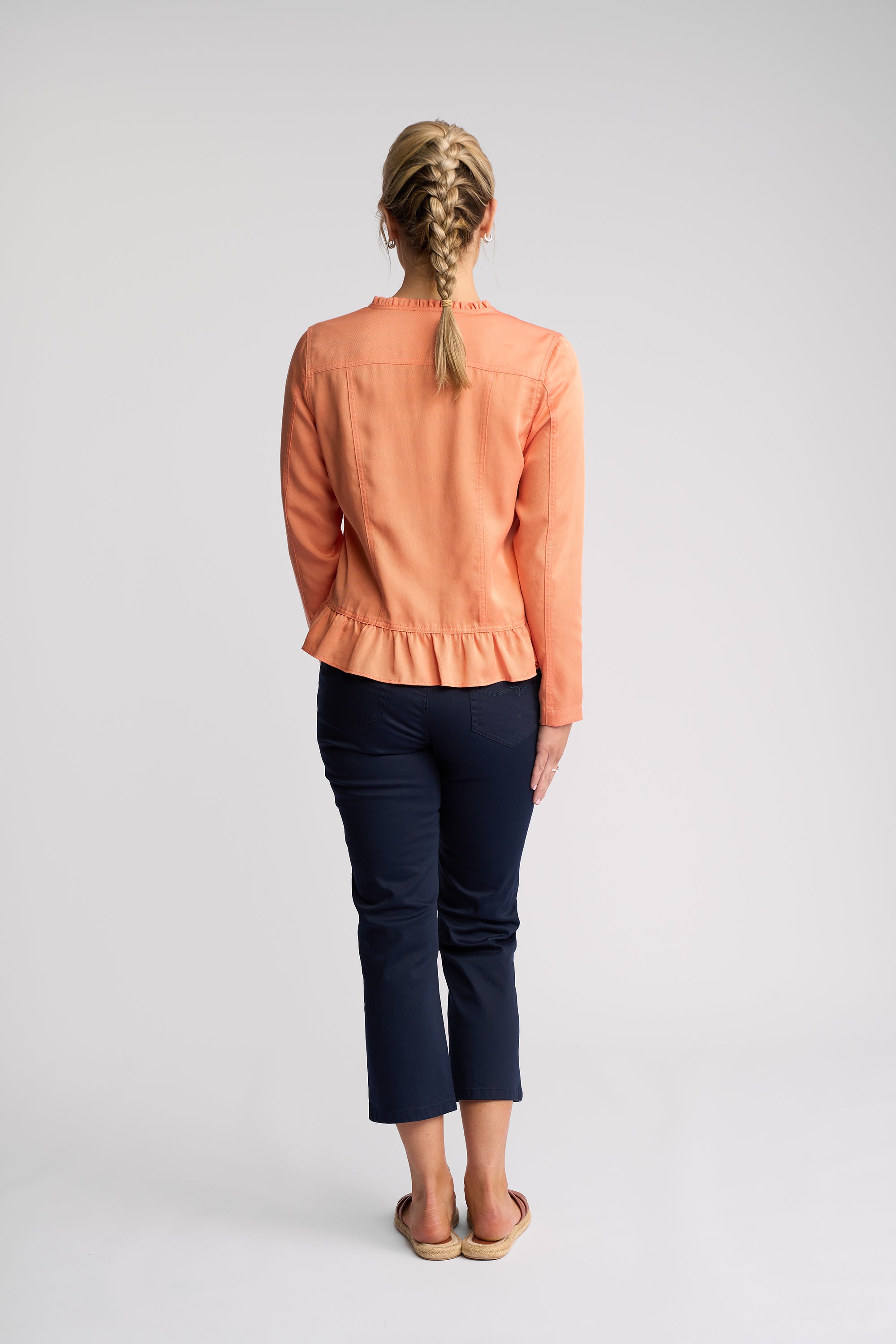 A woman viewed from behind wearing a peach-colored long-sleeve jacket with a ruffled hem and dark navy cropped pants, standing against a light gray studio background.