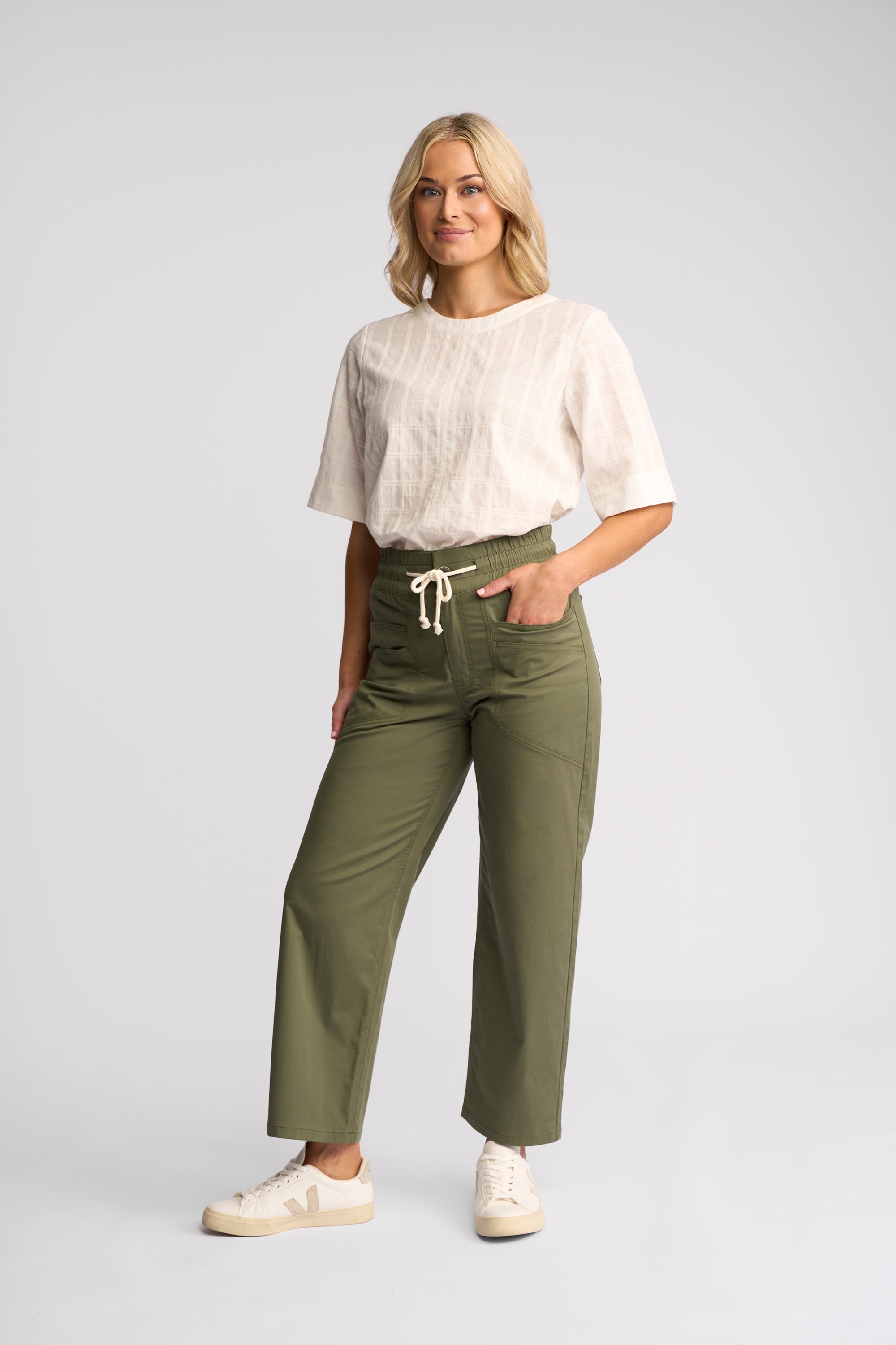 A woman modeling olive green wide-leg cropped pants with a cream drawstring waist, paired with a white textured short-sleeve top and white sneakers, against a light gray studio background.