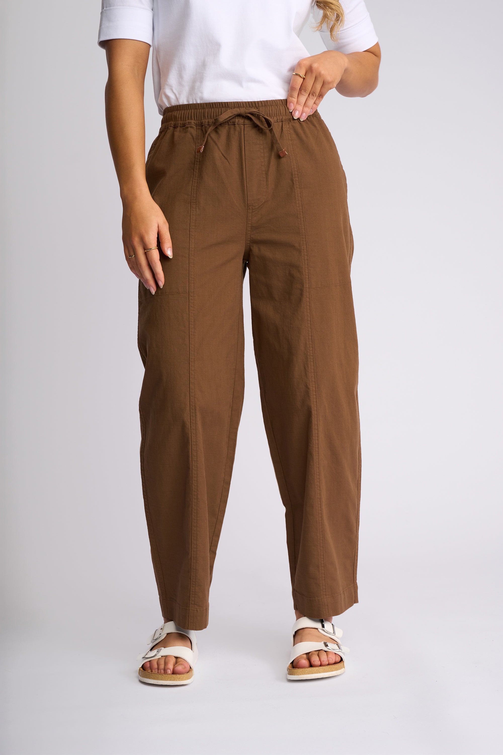 A person wearing brown drawstring wide-leg pants with a white t-shirt and white slide sandals, shown from the waist down against a light gray studio background.