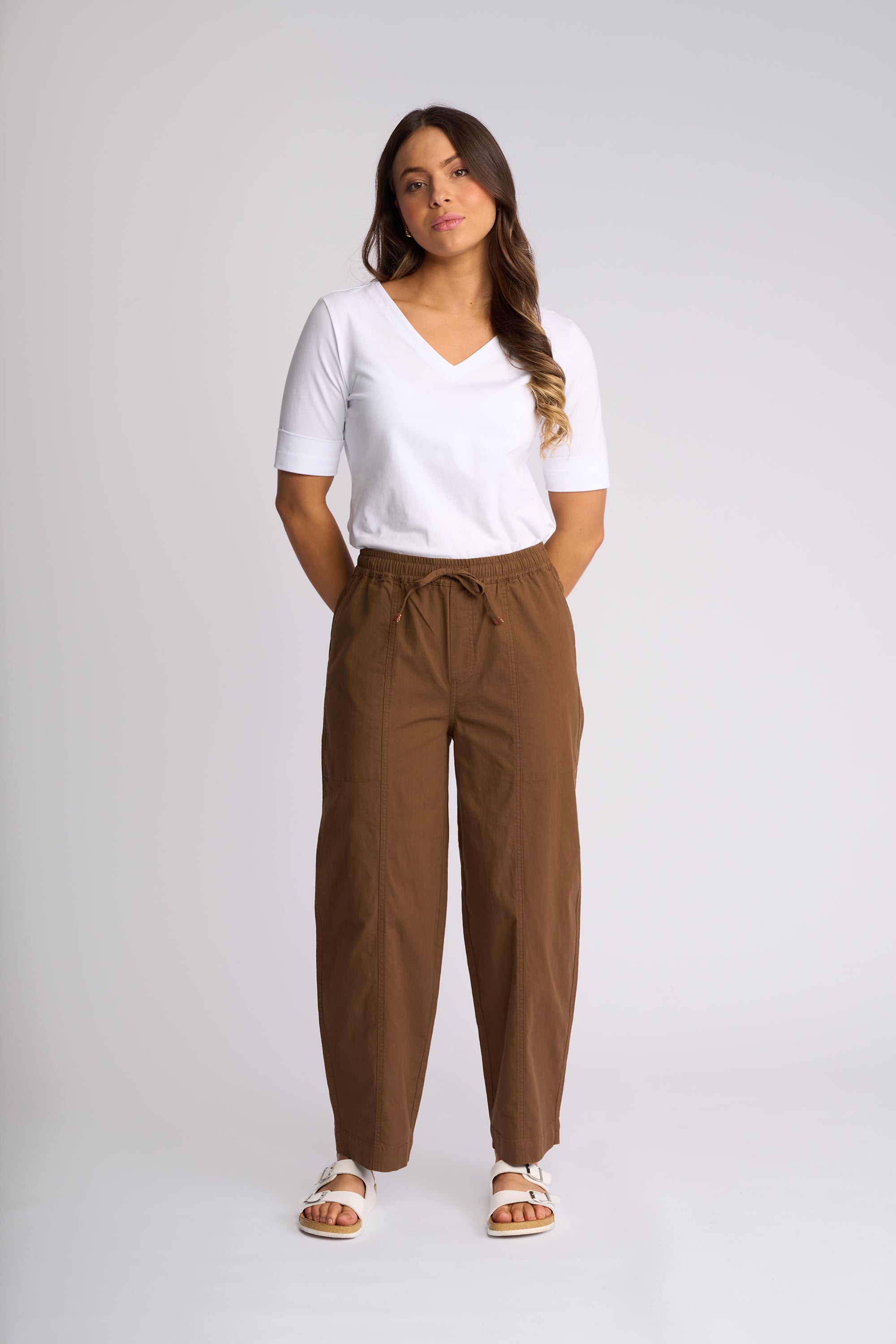 A woman modeling a pair of brown wide-leg drawstring pants, styled with a white V-neck t-shirt and white slide sandals, against a light gray studio background.