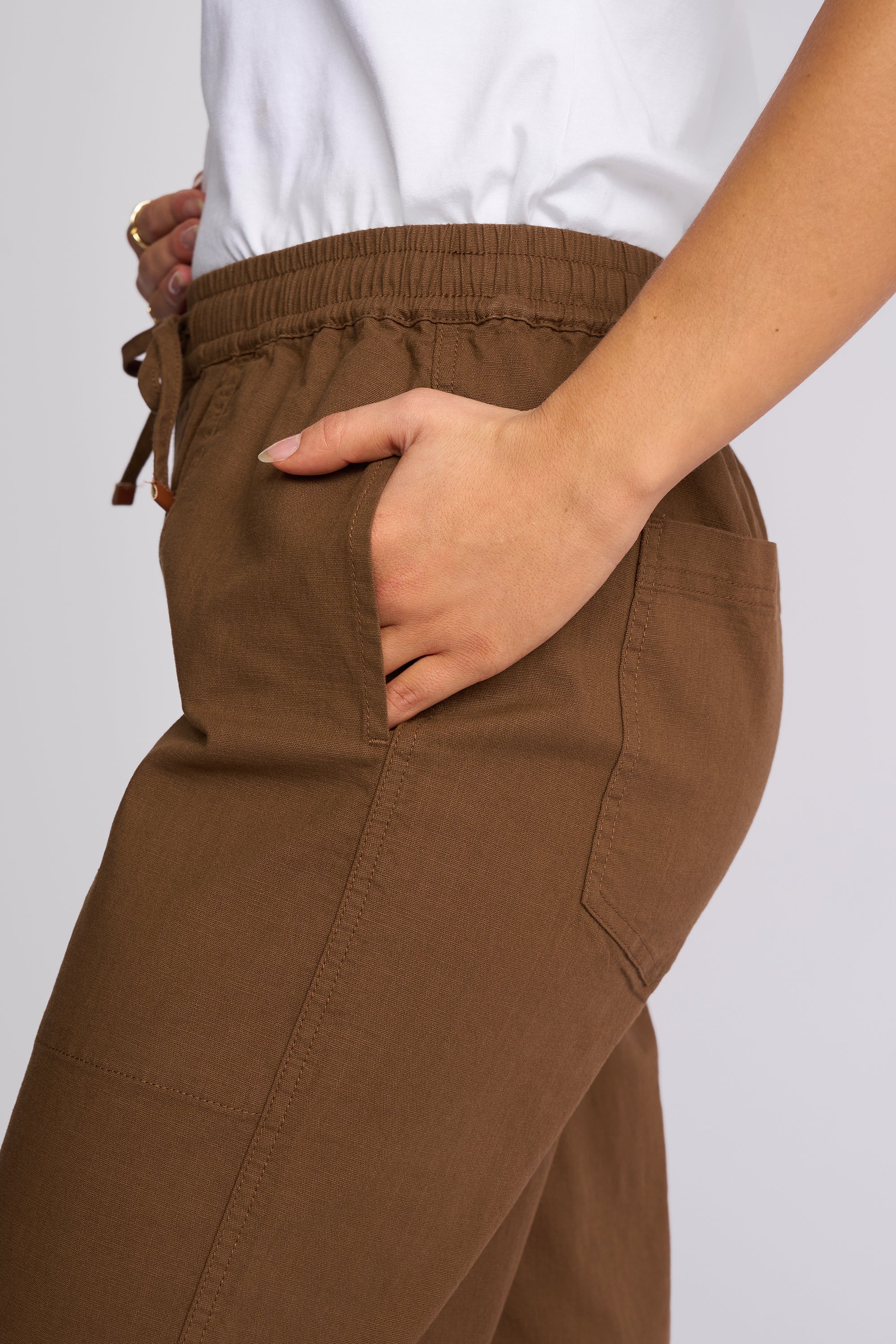 Close-up of a person wearing brown wide-leg pants with an elastic drawstring waist and side pockets, paired with a white top against a light gray background.