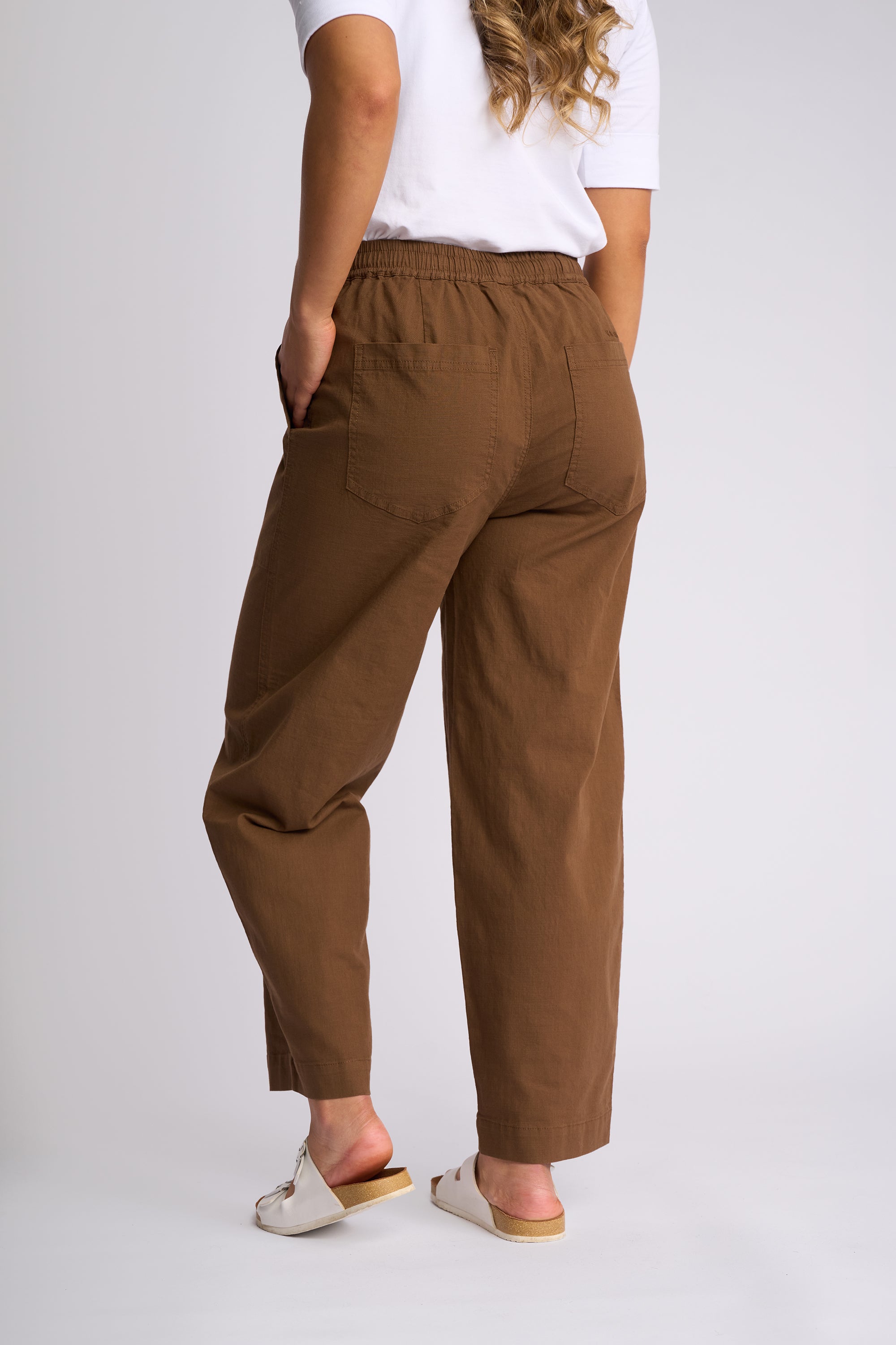 A person shown from the back wearing loose brown cropped pants with a white T-shirt and white slide sandals, photographed against a light gray studio background.