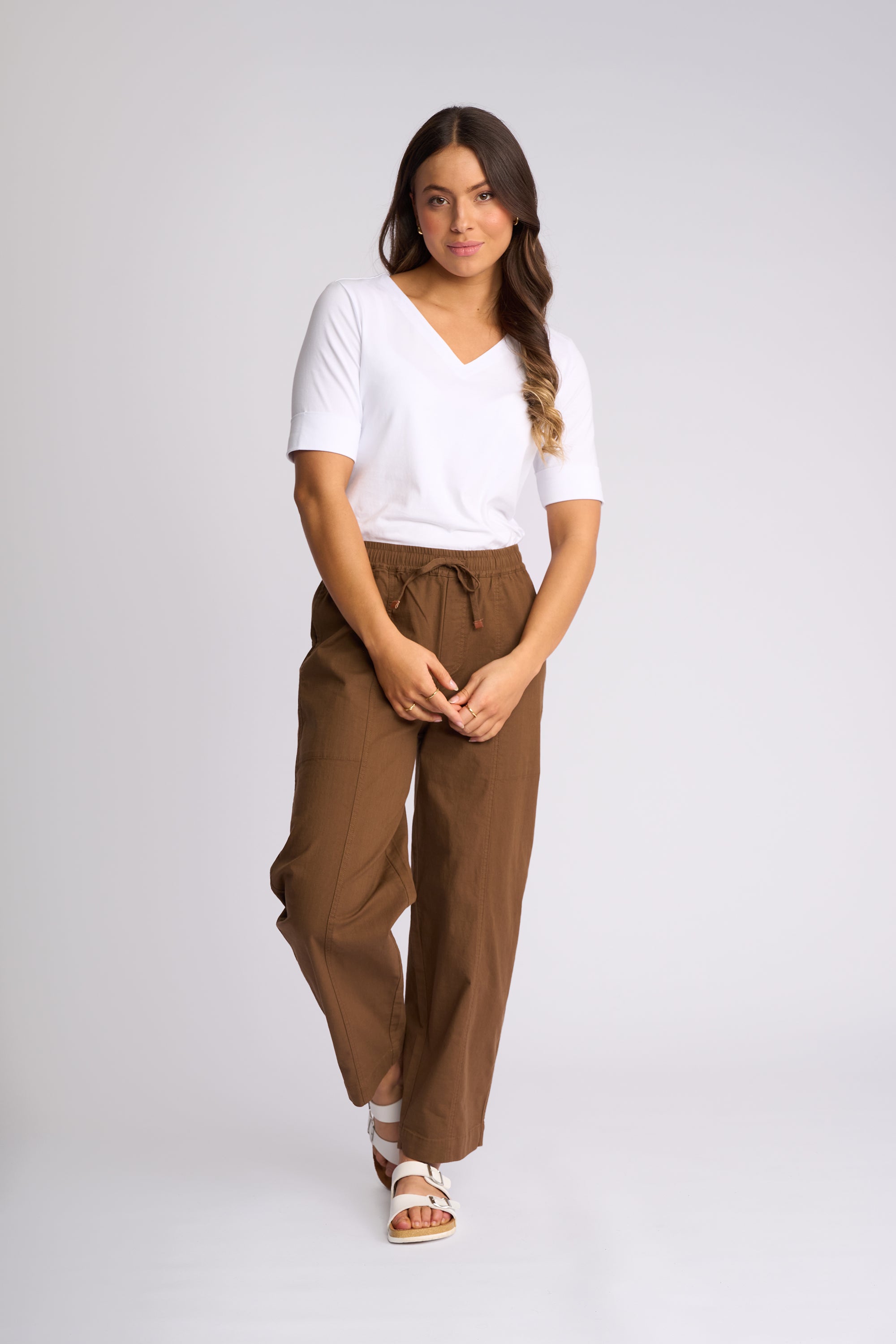 A woman modeling a white short-sleeve V-neck top and brown wide-leg drawstring pants against a light gray studio background.