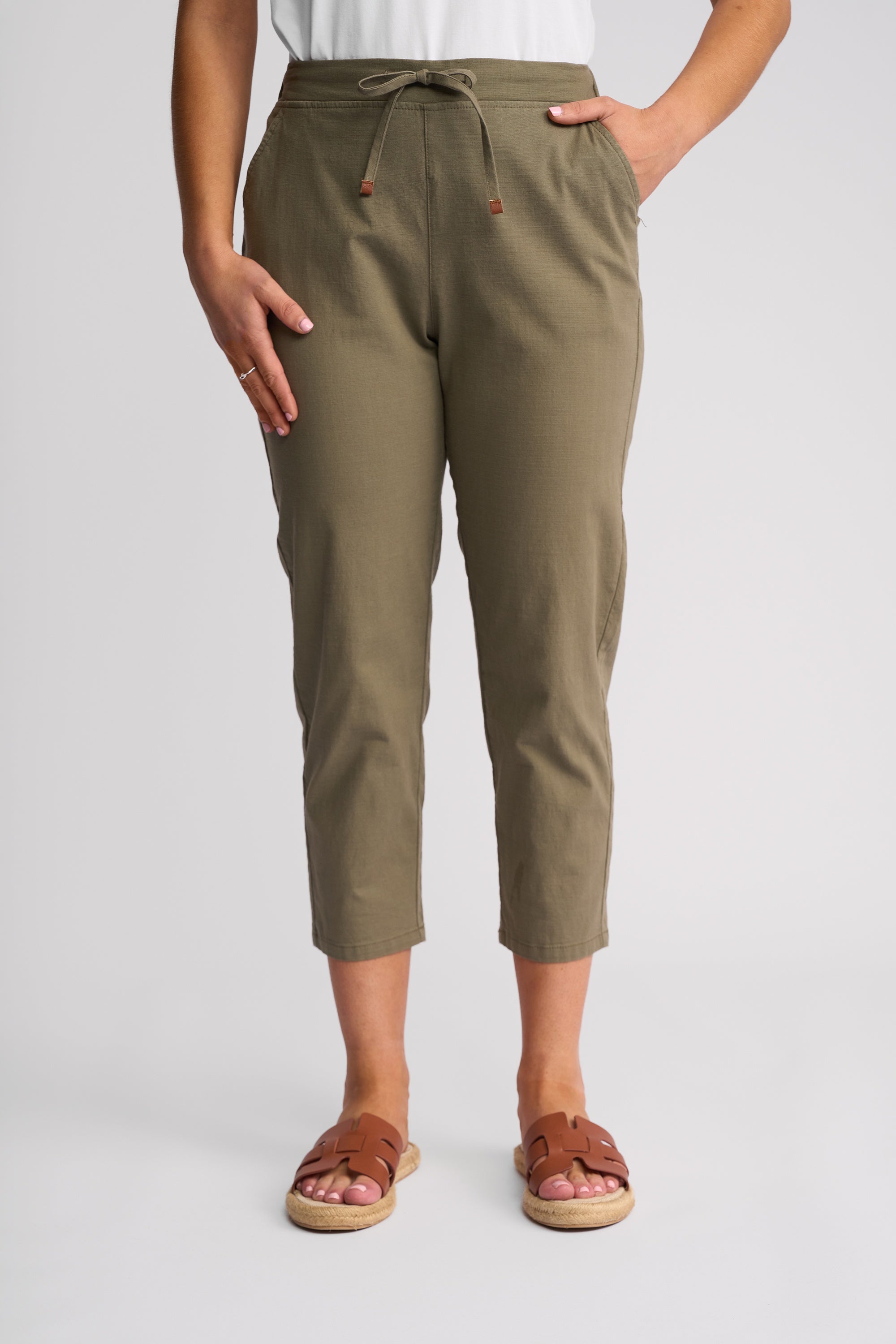 A person wearing olive green cropped drawstring pants with a white top and brown slide sandals, shown from the waist down against a light gray studio background.