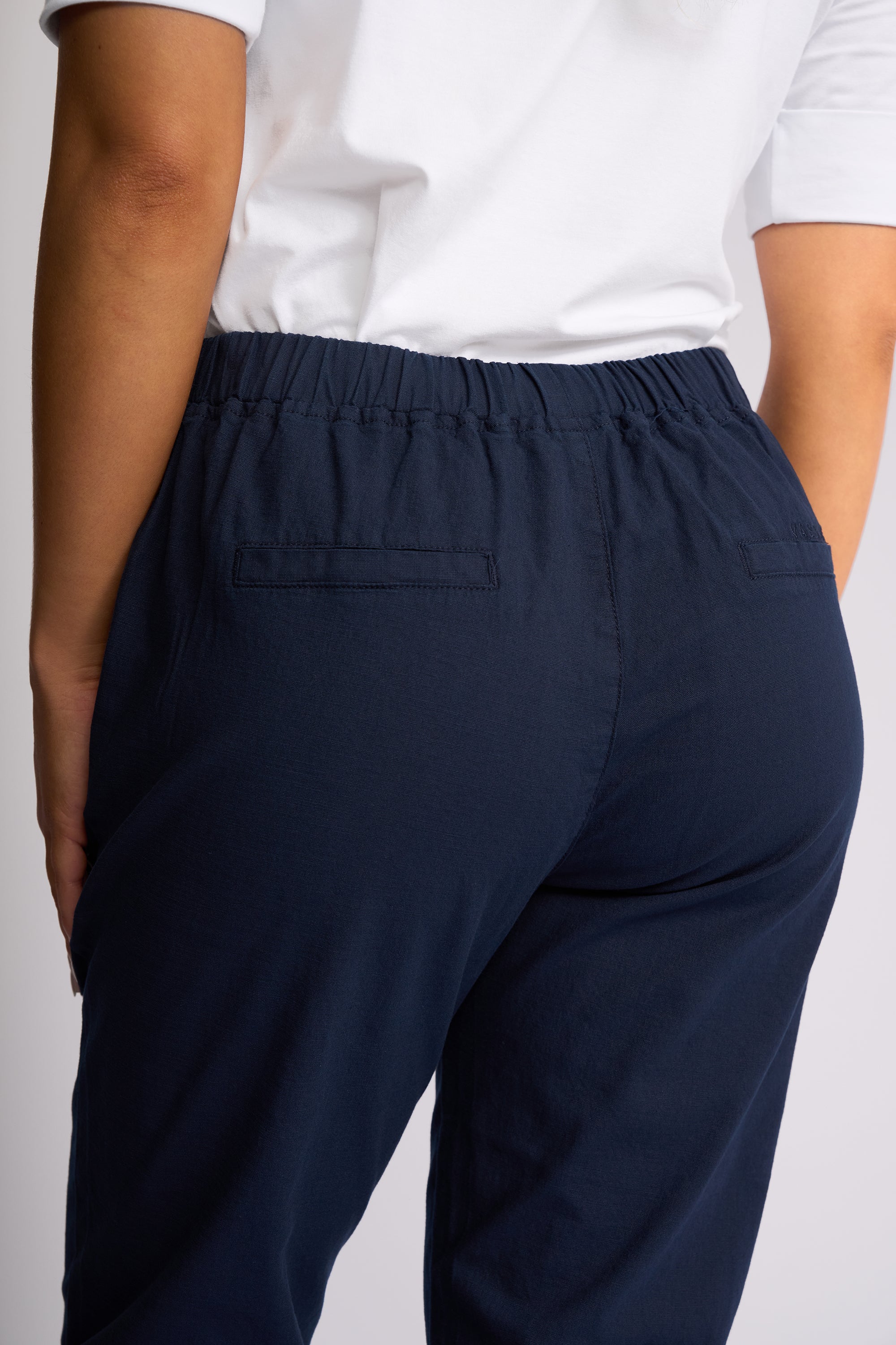 Rear view of a person wearing dark navy blue elastic-waist pants with back patch pockets, paired with a white top, against a light gray studio background.