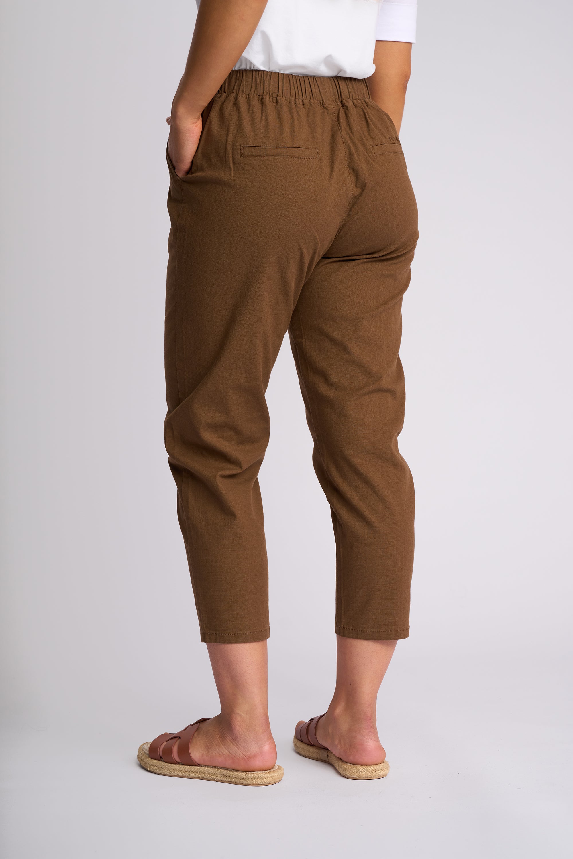 A person shown from the back wearing brown cropped pants with an elastic waistband, a white top, and brown slide sandals against a light gray studio background.
