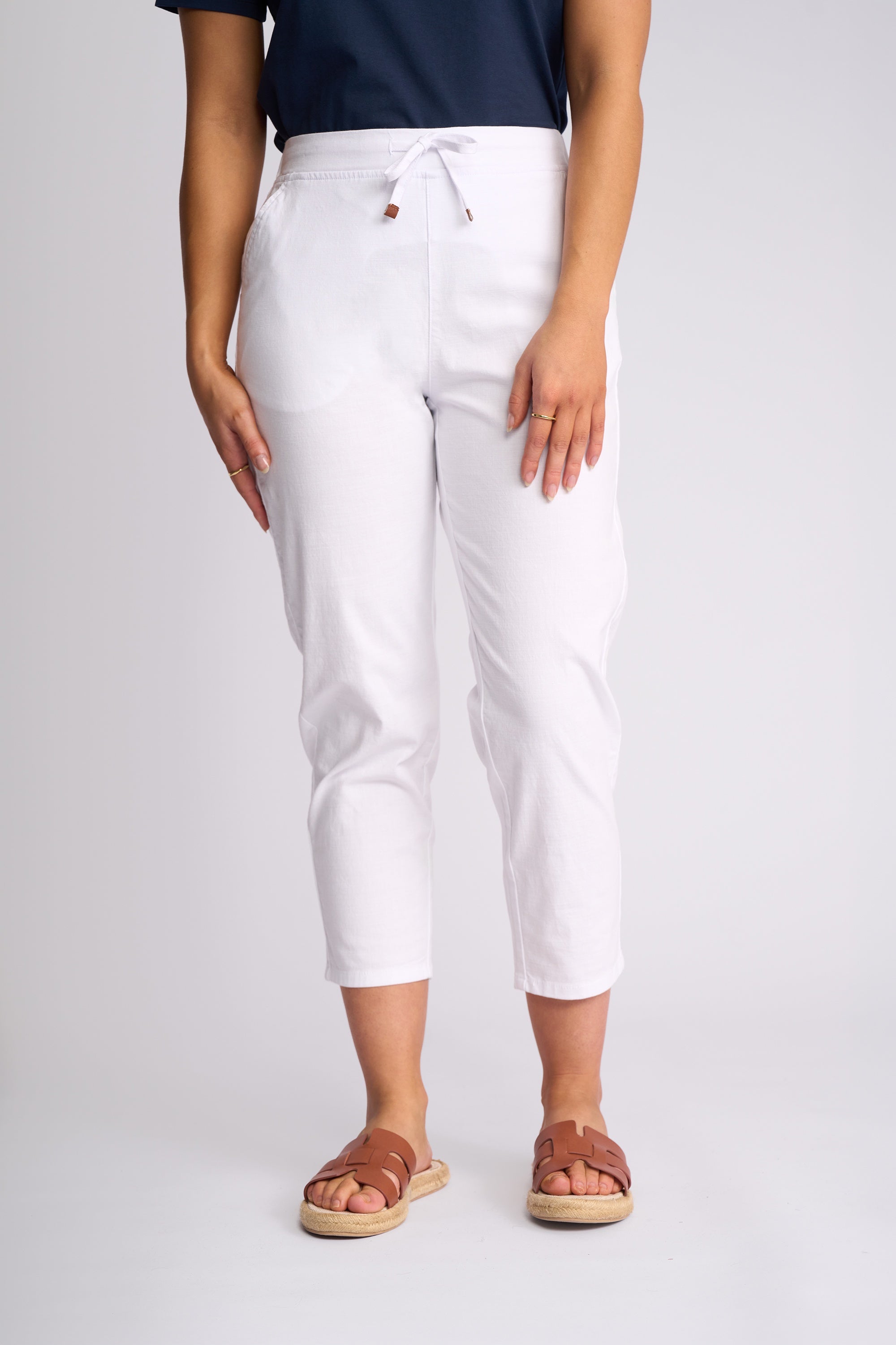 White cropped drawstring pants worn by a model, paired with a navy top and brown slide sandals, photographed against a light gray studio background.
