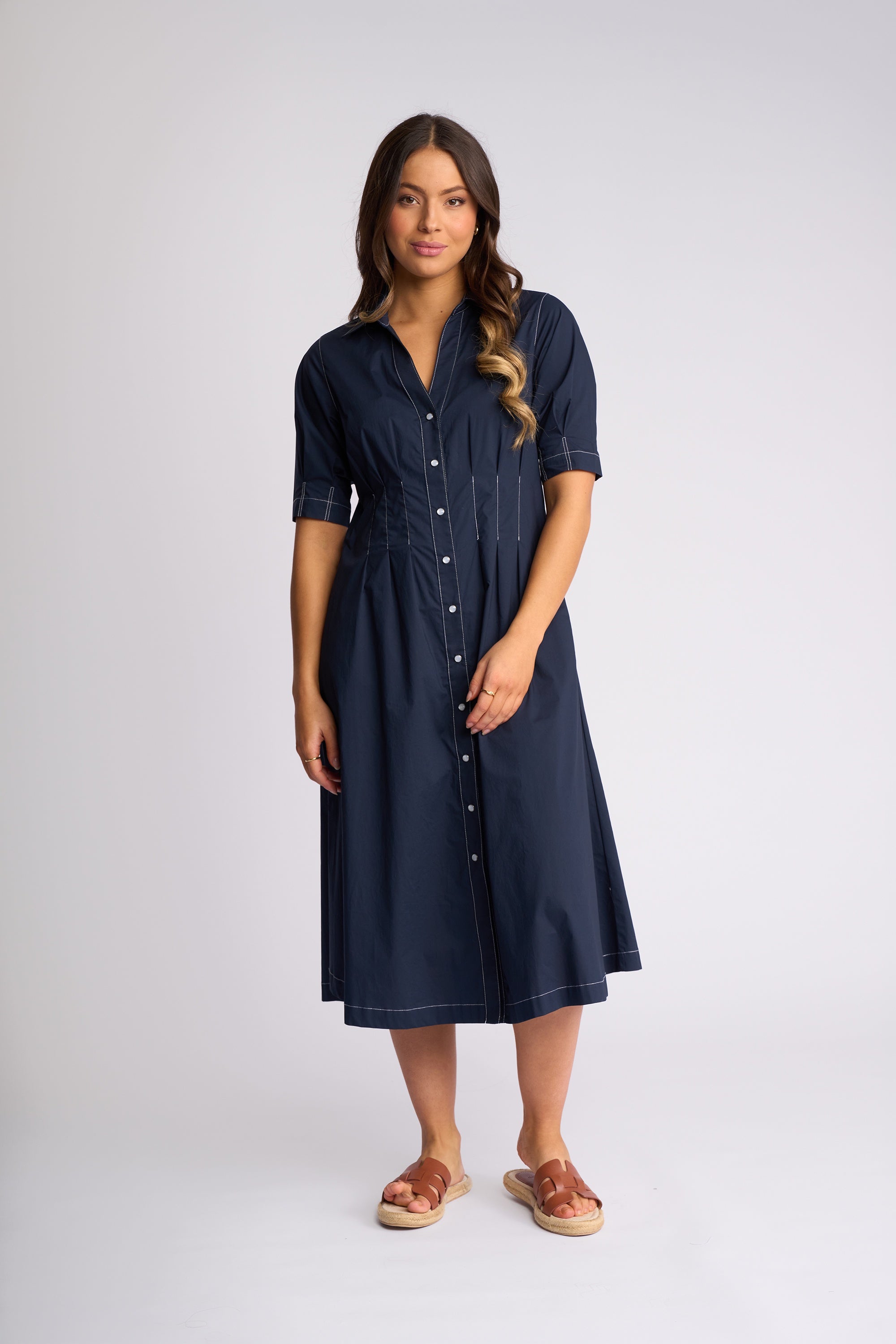 A woman modeling a dark navy blue midi shirt dress with short sleeves, a button-down front, and a gathered waist, paired with brown espadrille sandals against a light gray studio background.