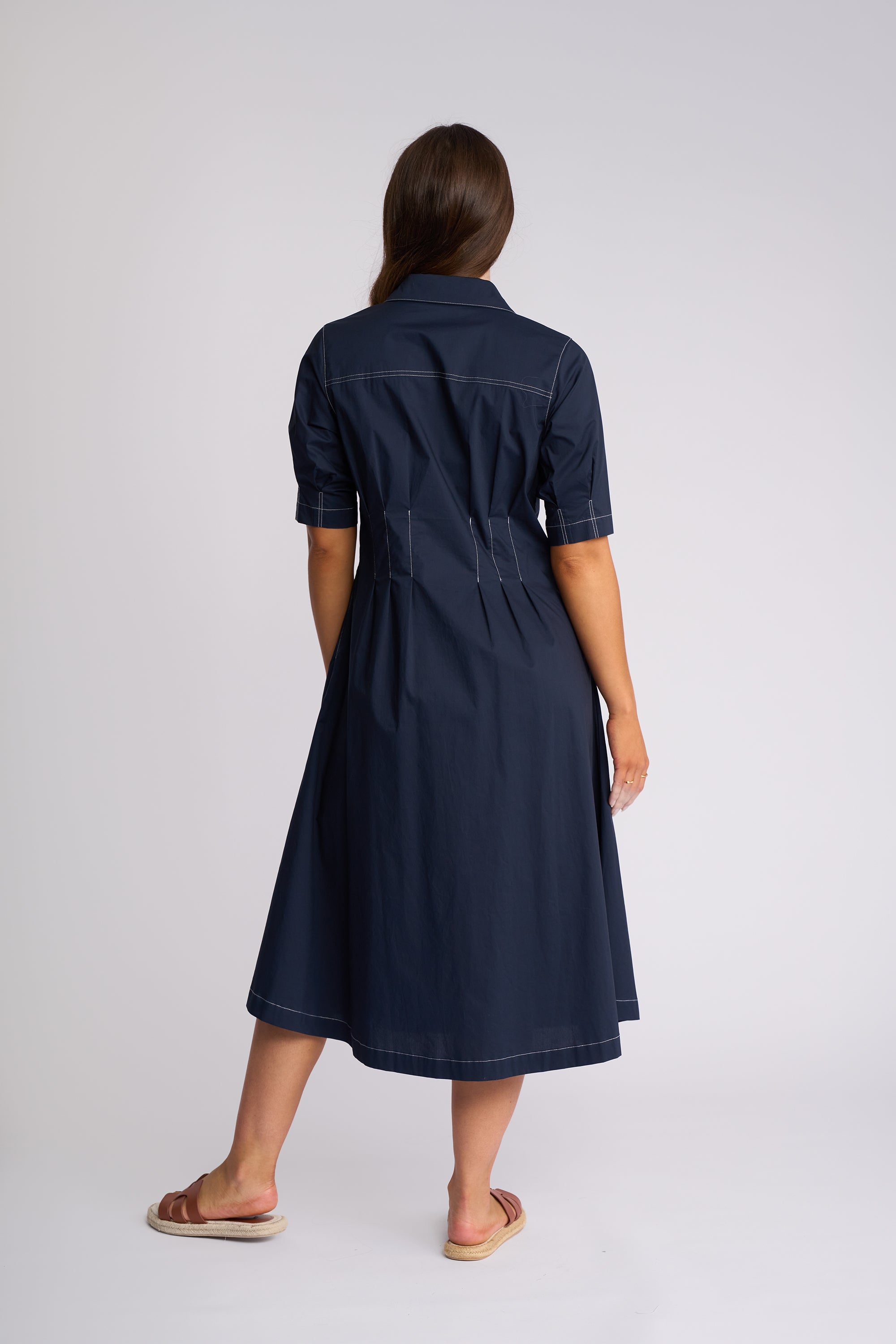 A woman viewed from the back wearing a dark navy midi shirt dress with short sleeves, a collar, and a gathered waist seam, paired with tan slide sandals against a light gray studio background.