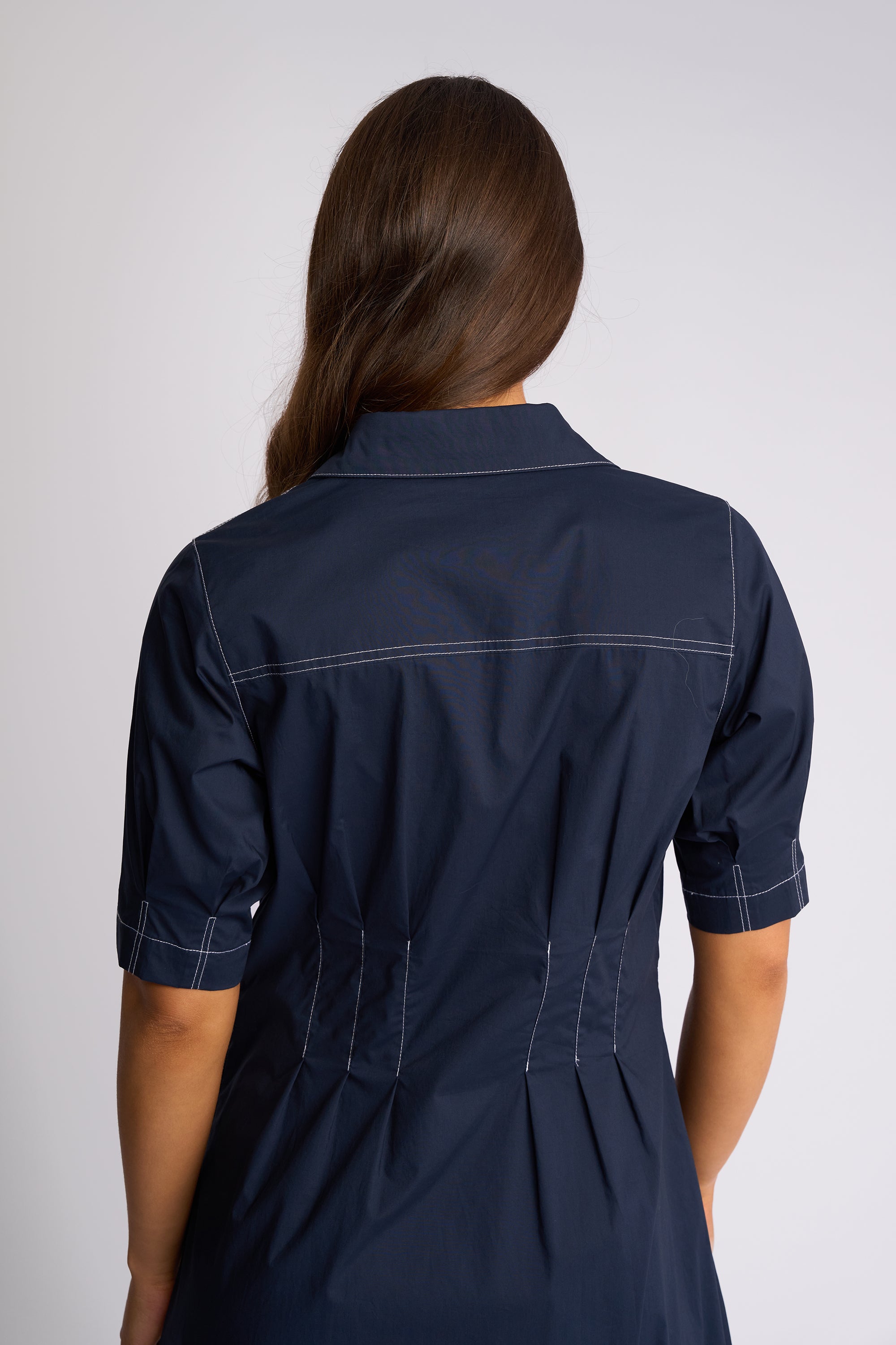 A woman viewed from the back wearing a navy blue short-sleeve shirt dress with white contrast stitching, set against a plain light gray studio background.