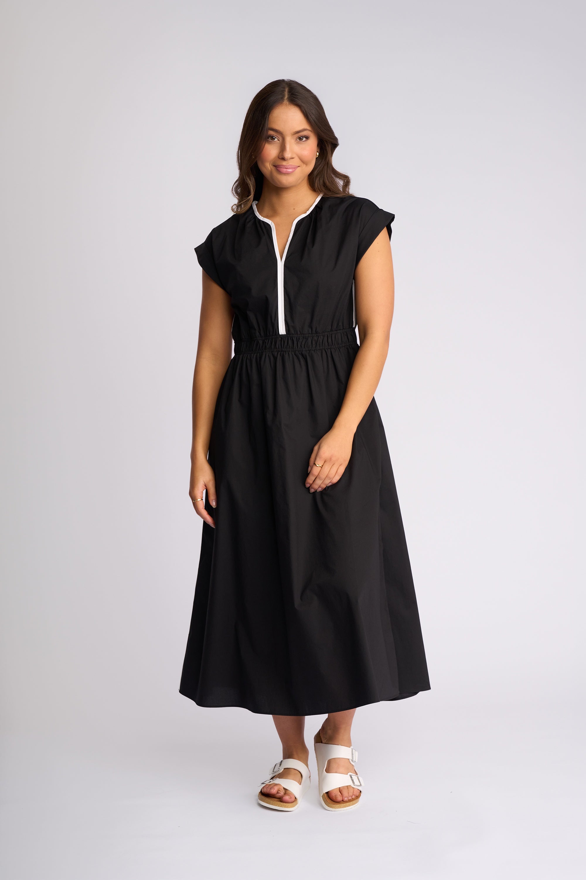 A woman modeling a black midi dress with a white zipper detail at the neckline, standing against a light gray studio background.