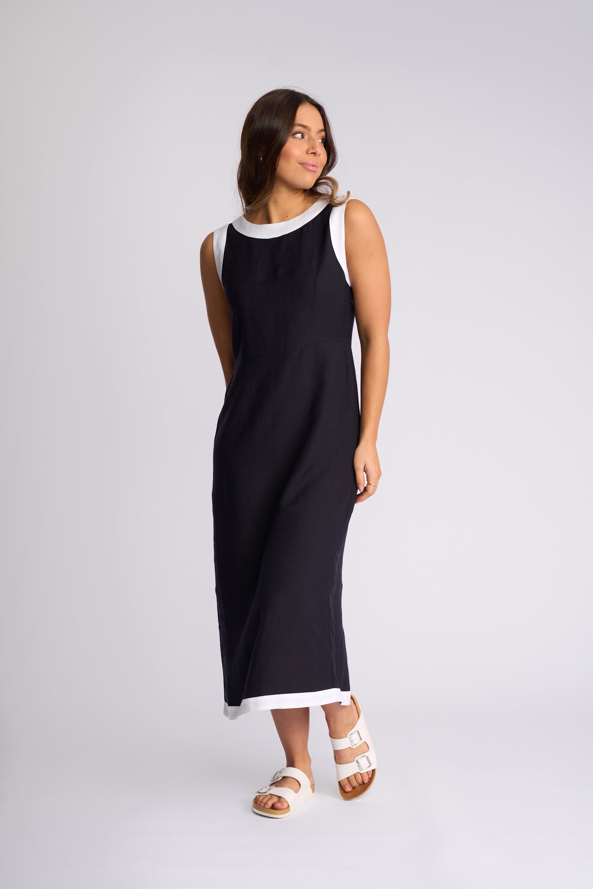A woman modeling a sleeveless black maxi dress with contrasting white trim at the crew neckline and hem, paired with white sandals, against a light gray studio background.