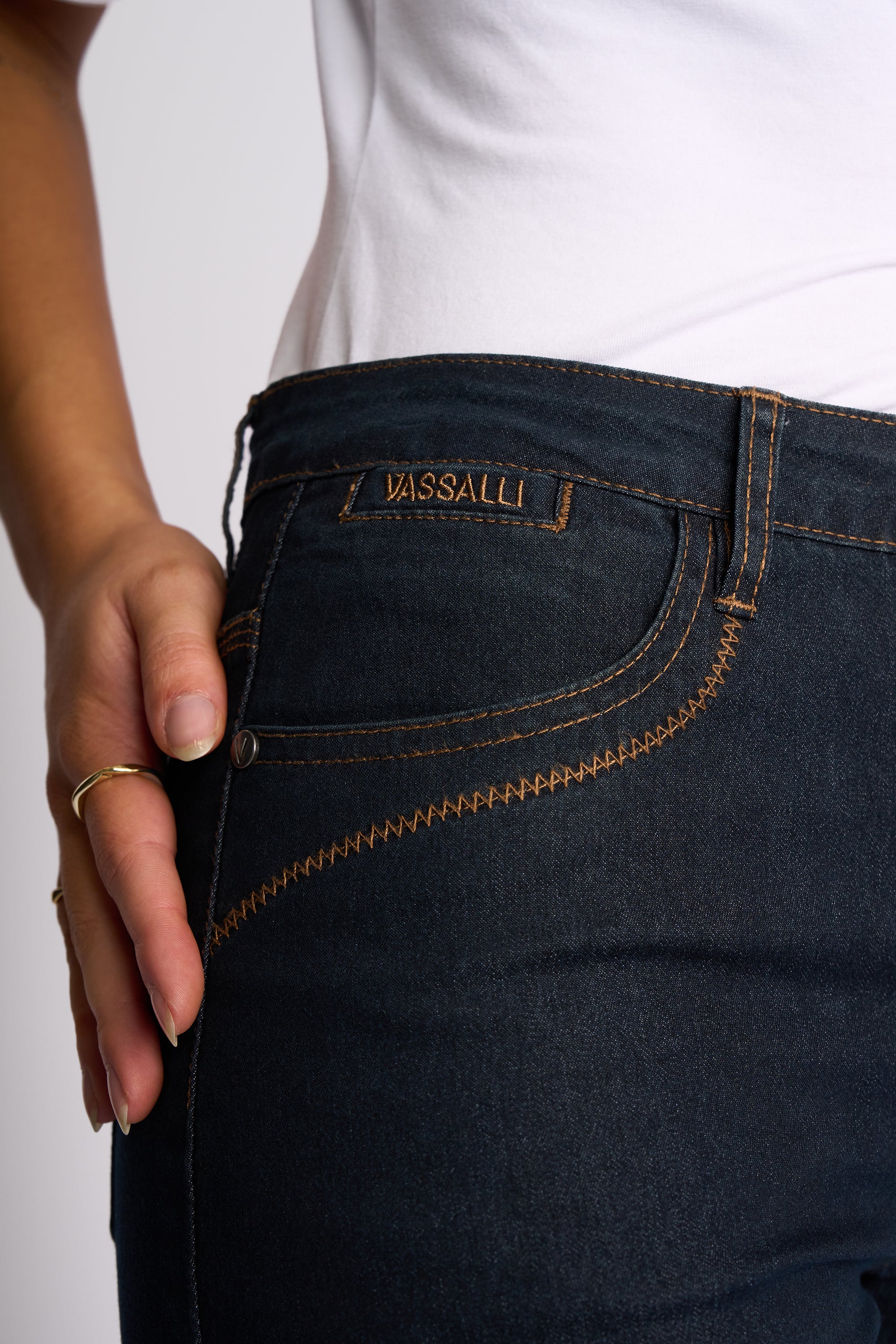 Close-up of a person wearing dark wash denim jeans with visible contrast stitching and a waistband patch reading PASSALLI. The person is also wearing a white top, and a hand with a gold ring rests near the pocket.